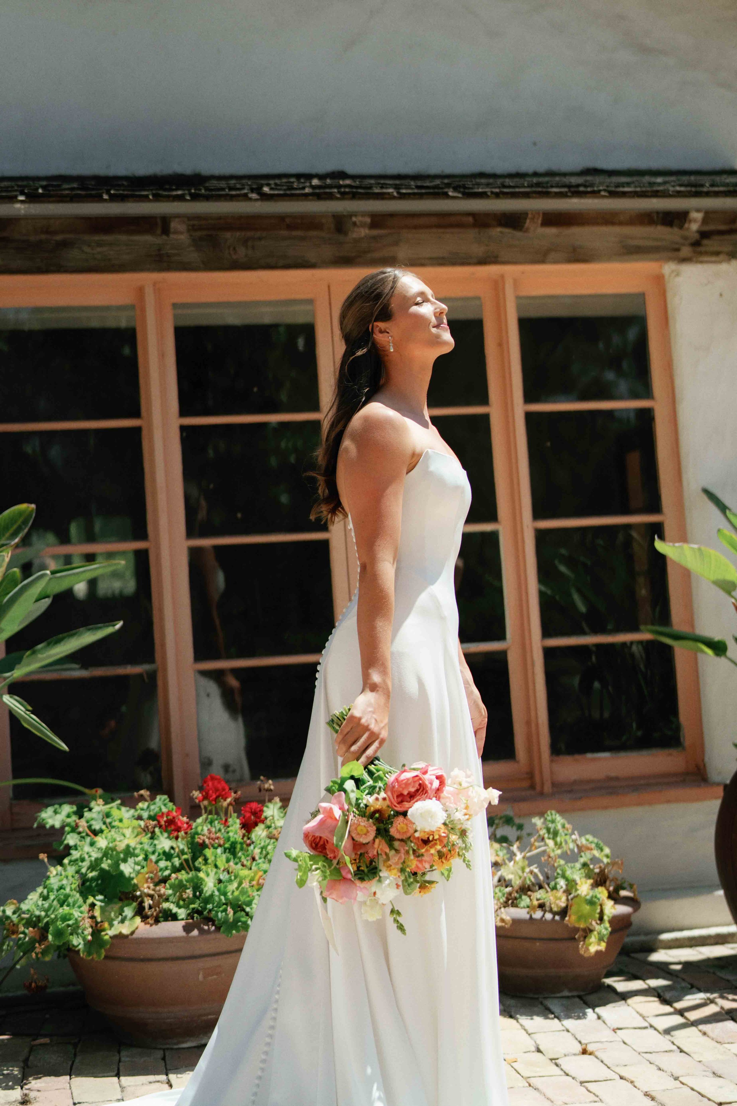 A woman in a strapless white wedding gown holding a large bouquet of pink and white flowers, standing outdoors in front of a building with large windows and potted plants, smiling with her eyes closed.