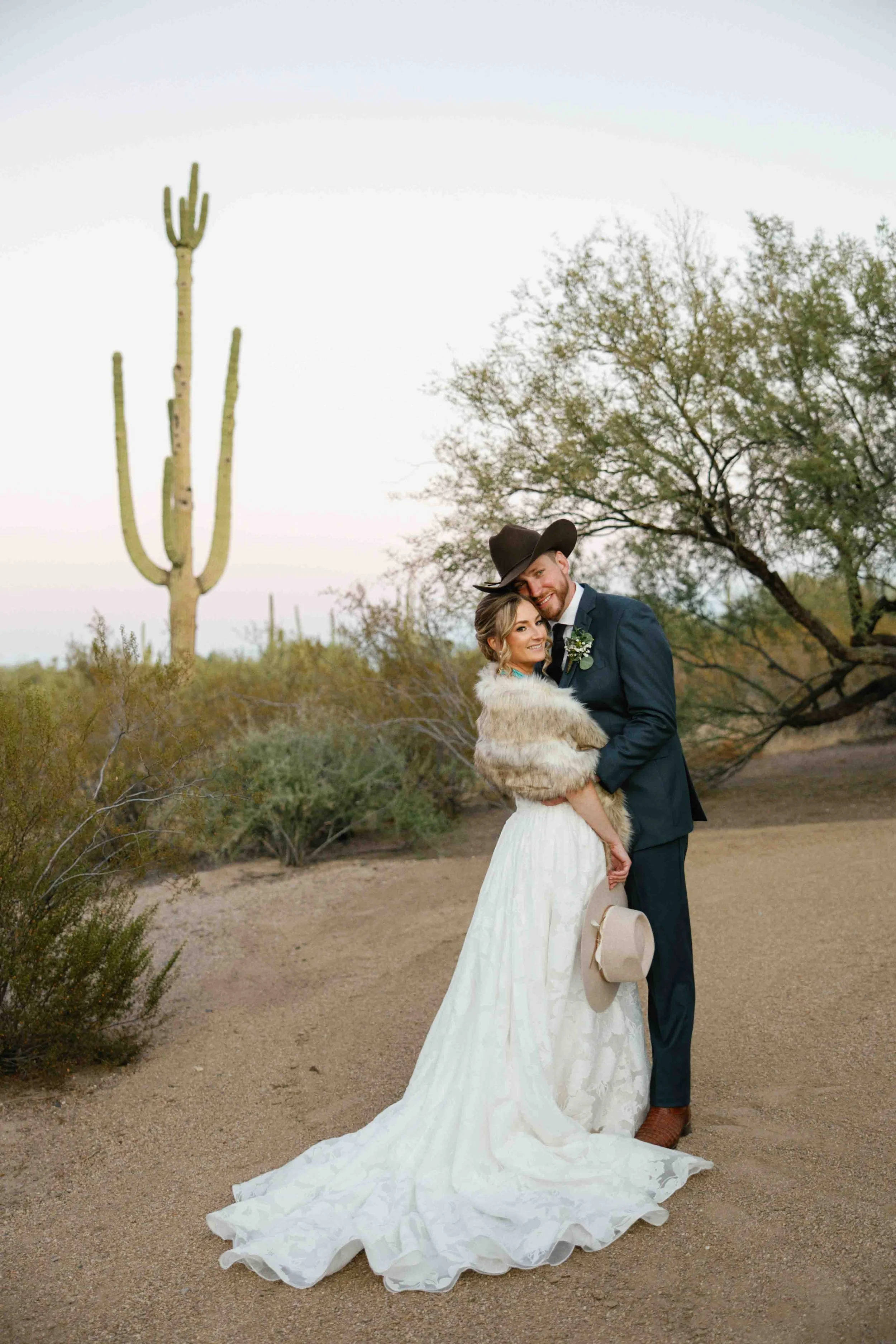 A newlywed couple dressed in wedding attire standing outdoors in a desert landscape with cacti and sparse trees. The bride is wearing a white wedding dress with a train and a faux fur shawl, and the groom is wearing a dark suit with a boutonniere and