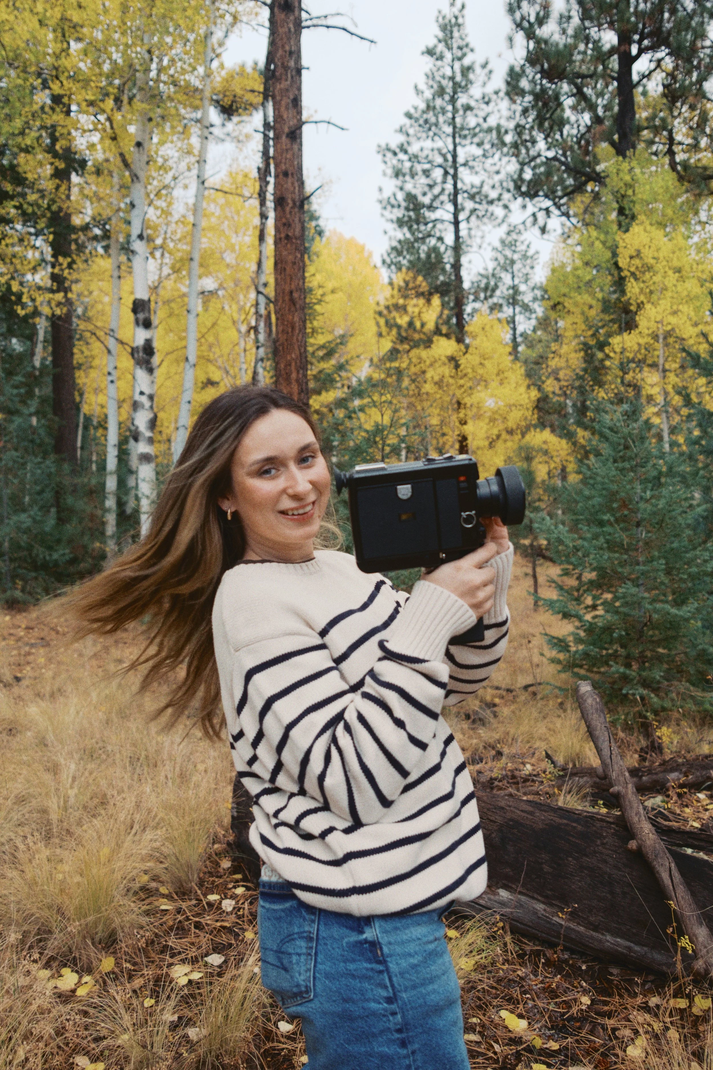 A young woman with long hair in a striped sweater and jeans outdoors, holding a camera in a forest with trees and fall foliage.