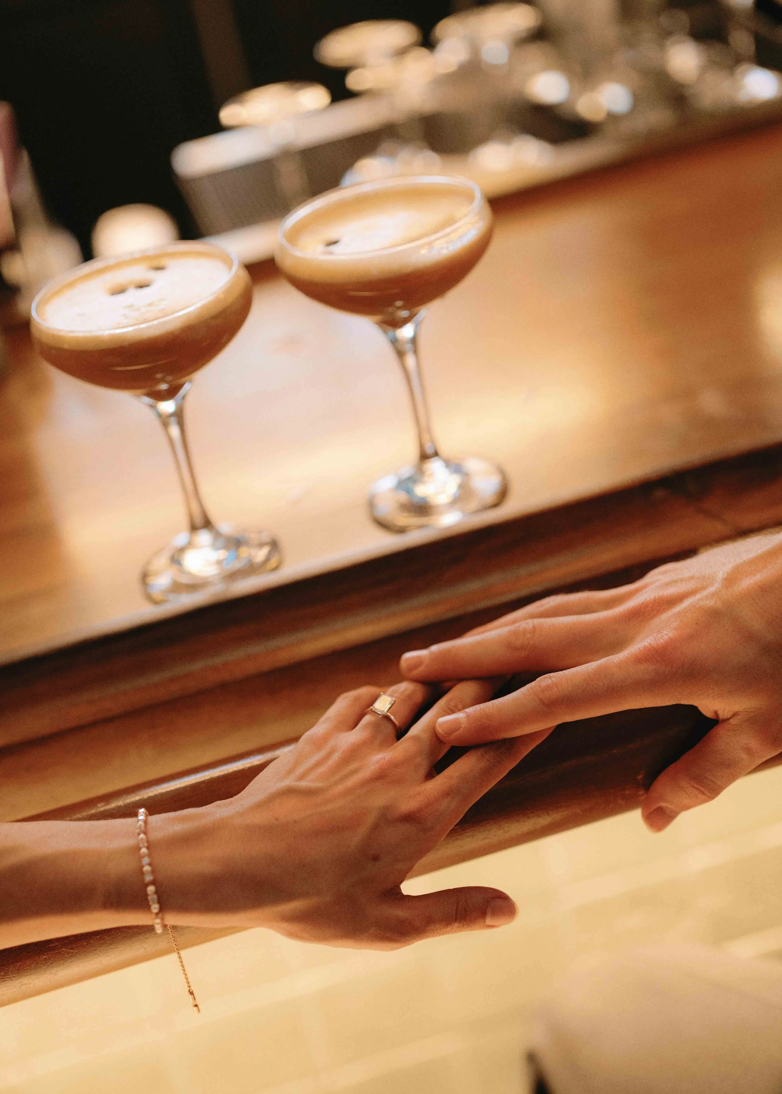 Two hands resting on a wooden surface with two glasses of coffee or espresso in the background and jewelry including a ring, bracelet, and necklace.
