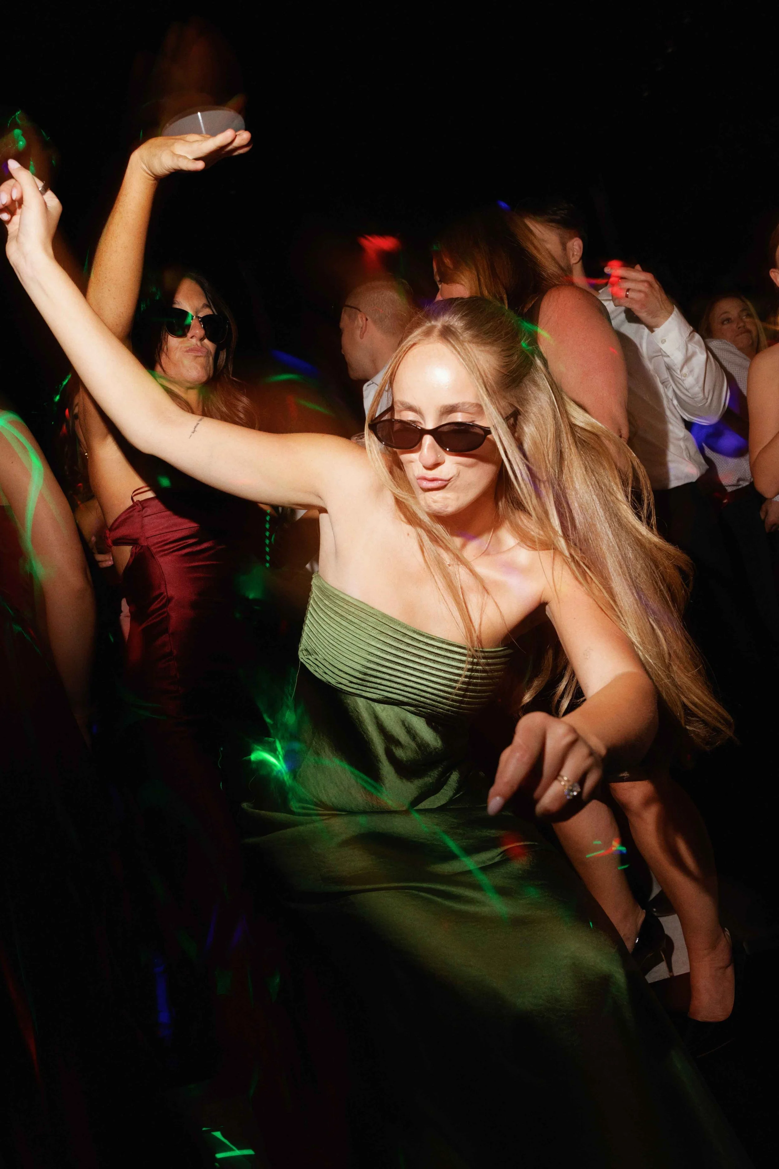 Group of people dancing at a party with colorful lights, some wearing sunglasses, enjoying the music and having fun.