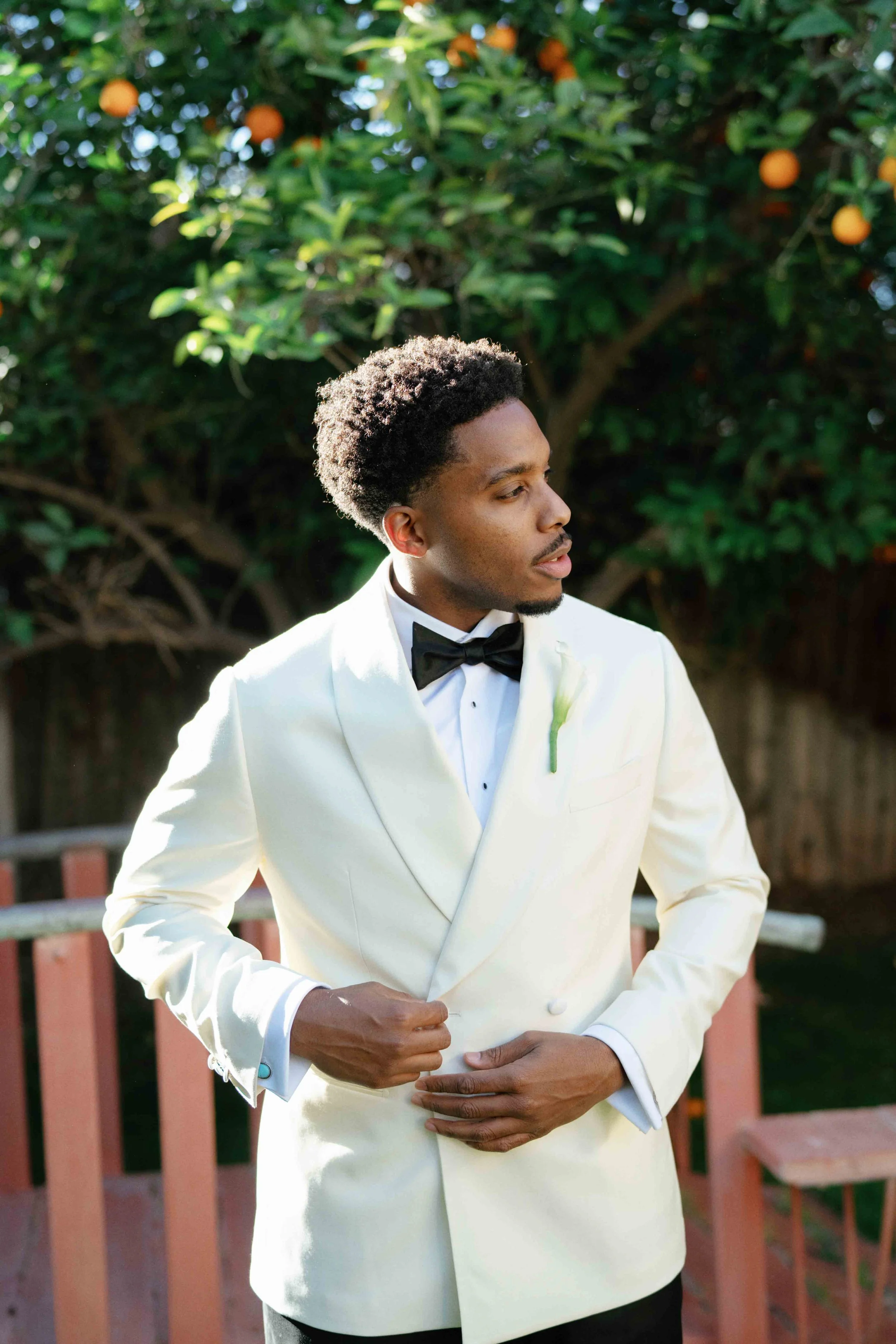 A man in a white tuxedo with a black bowtie and boutonniere, standing outdoors in front of a shaded orange tree, looking to the side with his hands folded.
