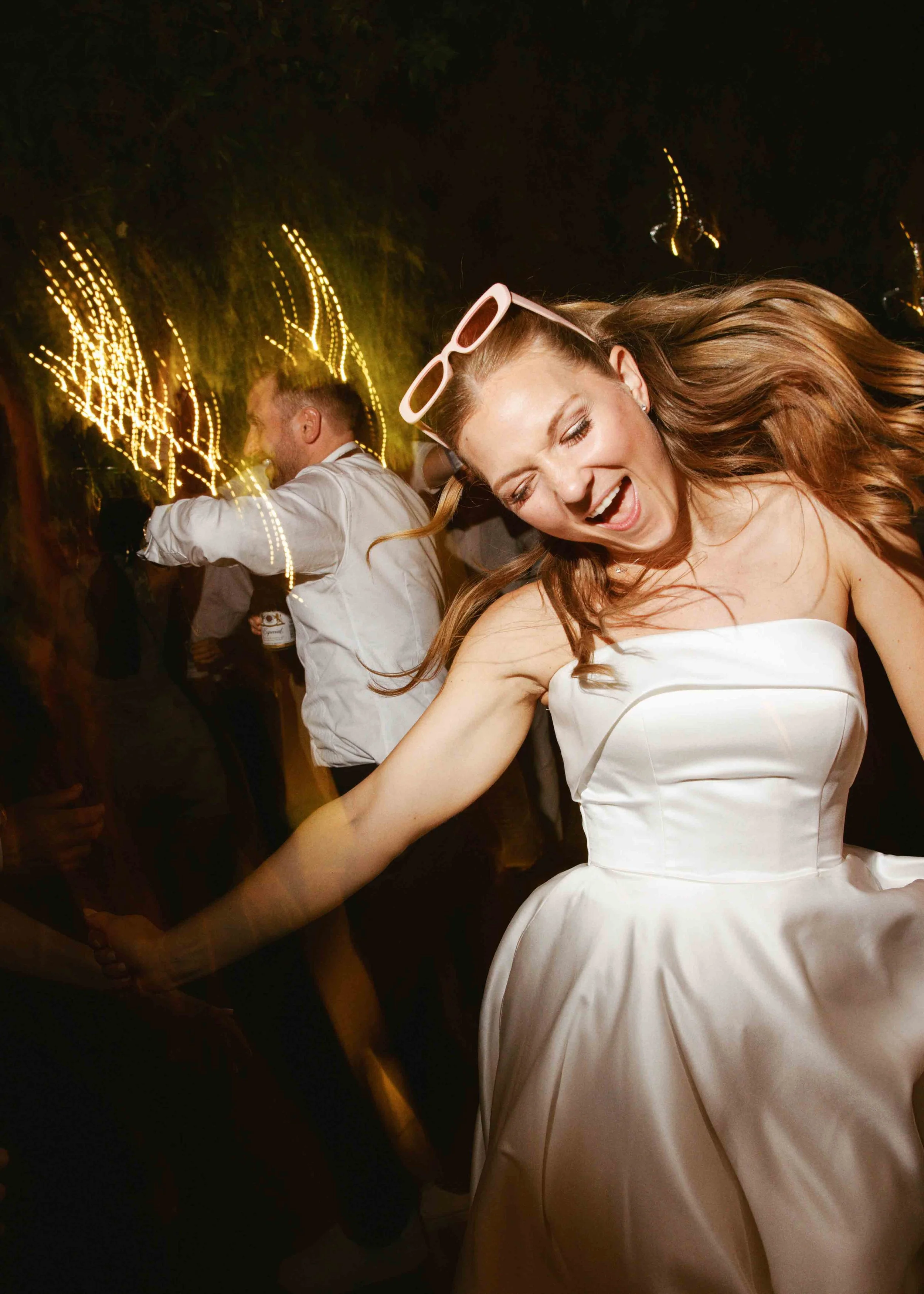 A woman in a strapless white dress dancing at a party with a big smile, wearing pink sunglasses on her head. A man in a light-colored shirt is dancing in the background with string lights creating a bokeh effect.
