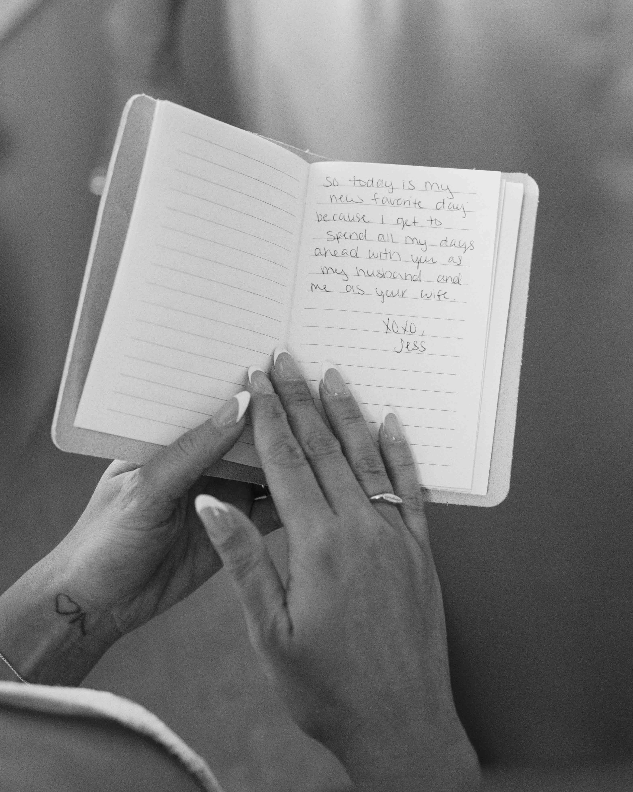 A person's left hand holding open a small notebook with handwritten text inside. The person has decorated nails and a tattoo on the wrist.