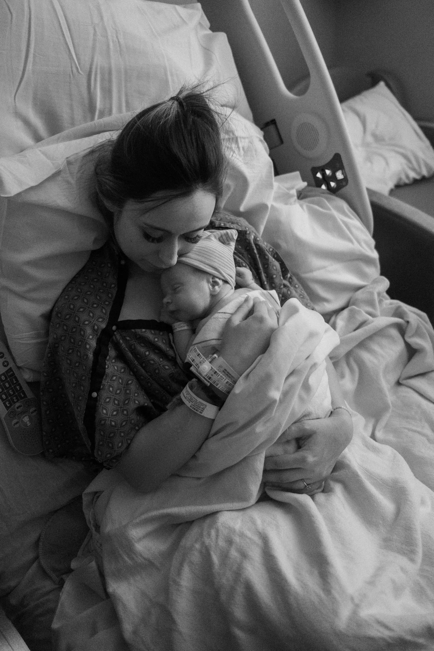 A woman in a hospital bed holding a sleeping newborn baby, with hospital wristbands and medical equipment visible.