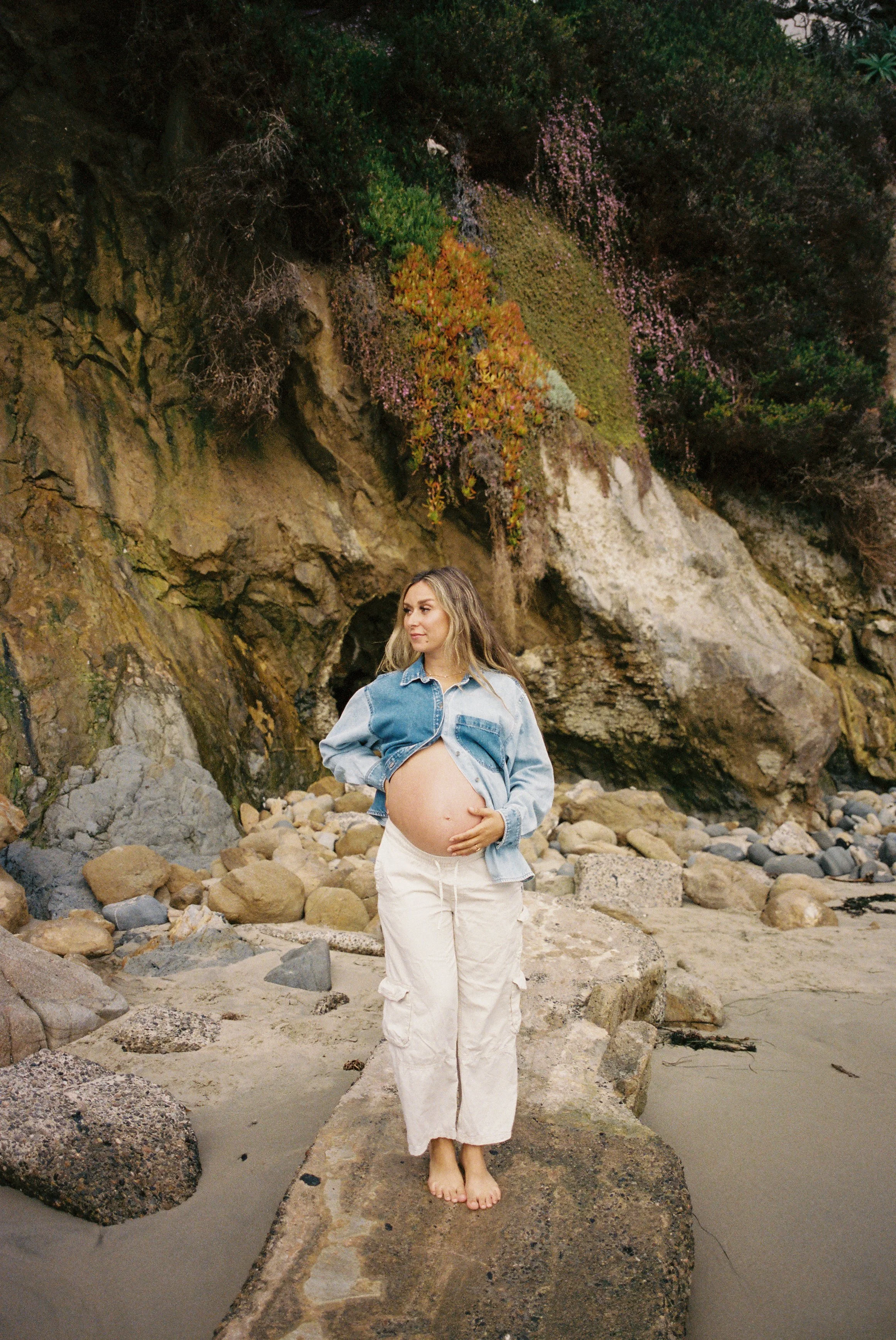 A pregnant woman standing barefoot on a large rock at the beach, holding her belly with one hand, with rocky terrain and a cliff covered in greenery and blooming pink flowers in the background.