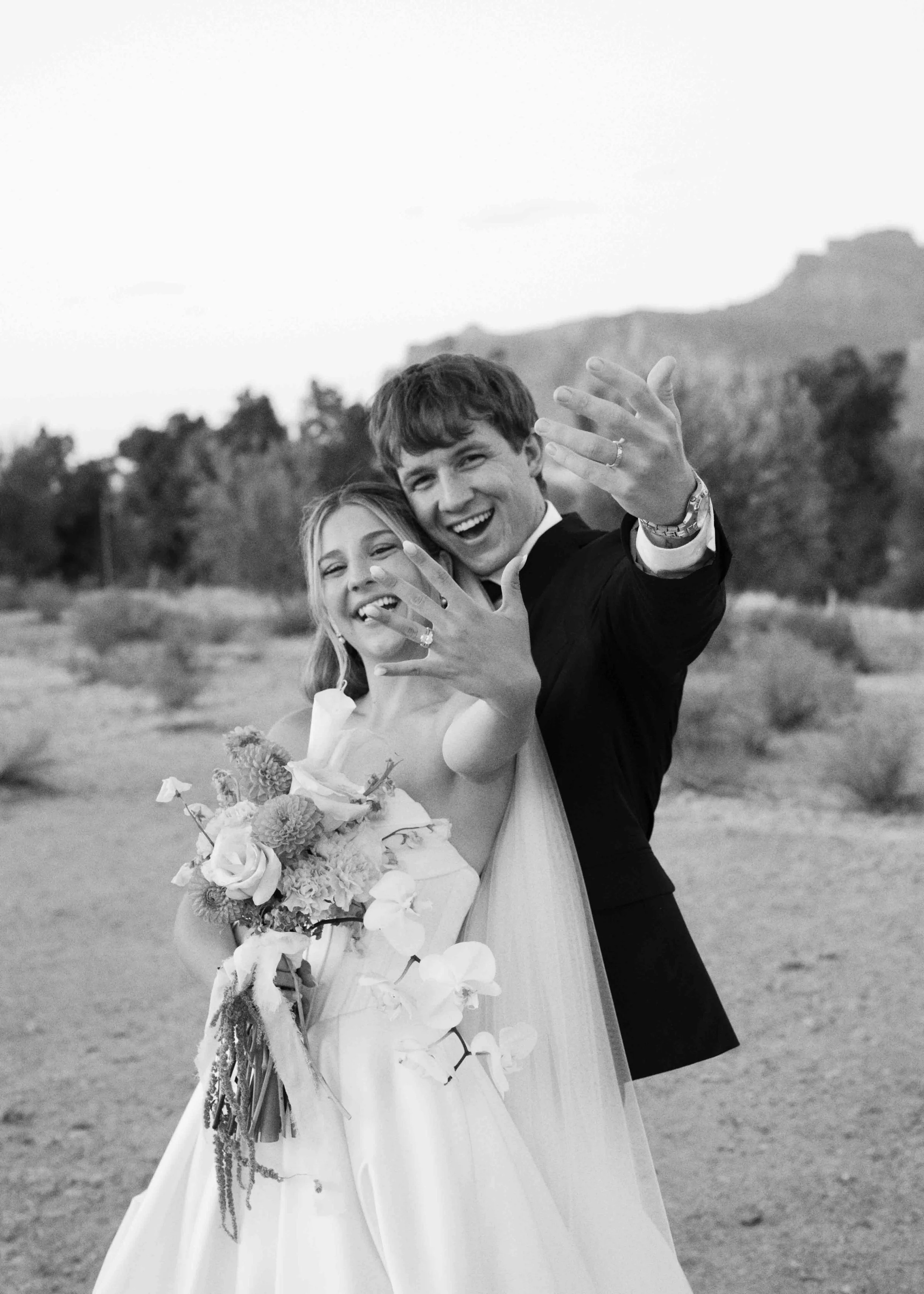 A smiling couple on their wedding day, the woman wearing a wedding dress and holding a bouquet of flowers, and the man in a tuxedo, outdoors with mountains and trees in the background.