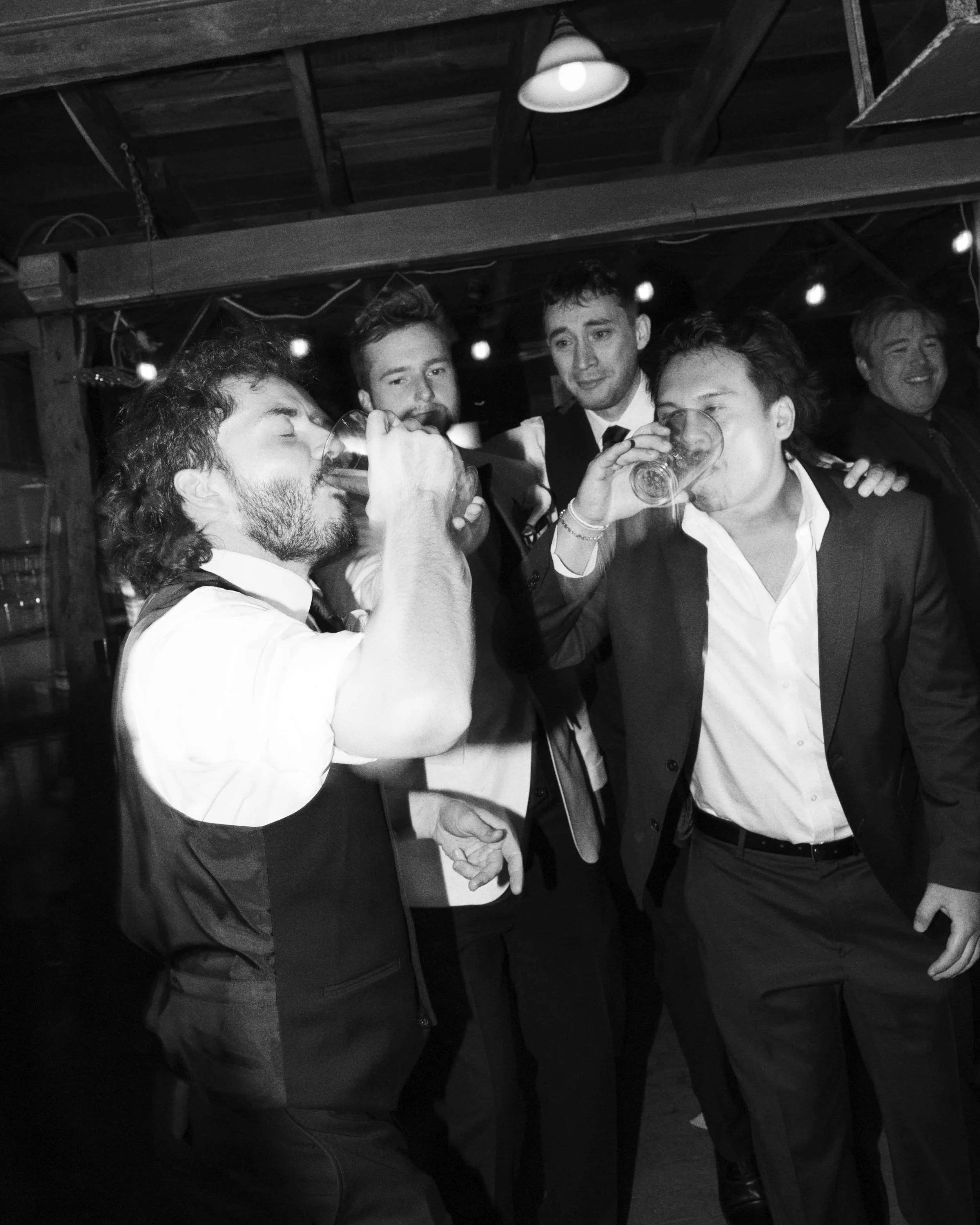 Group of five people in formal attire at a social gathering, some drinking from glasses and engaging in conversation, in a dimly lit indoor space with string lights and a wooden ceiling.