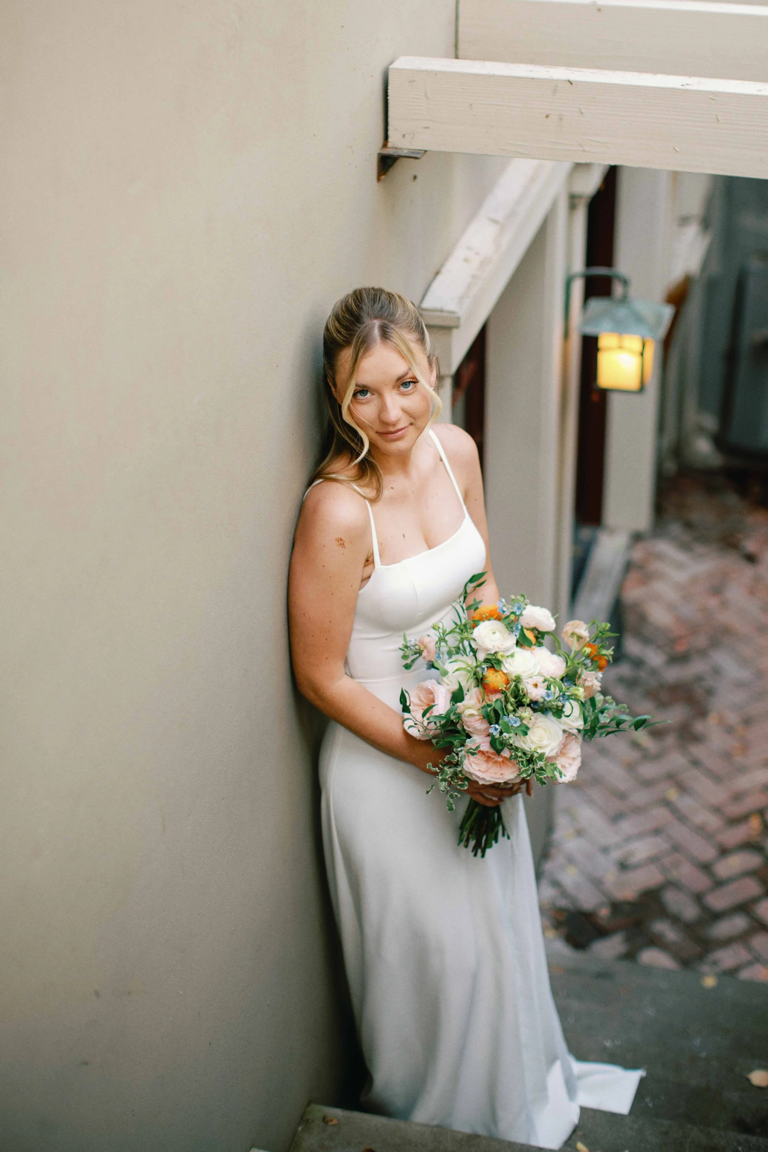 A bride with blonde hair and blue eyes in a white wedding dress is leaning against a beige wall, holding a bouquet of colorful flowers, and looking at the camera with a slight smile.