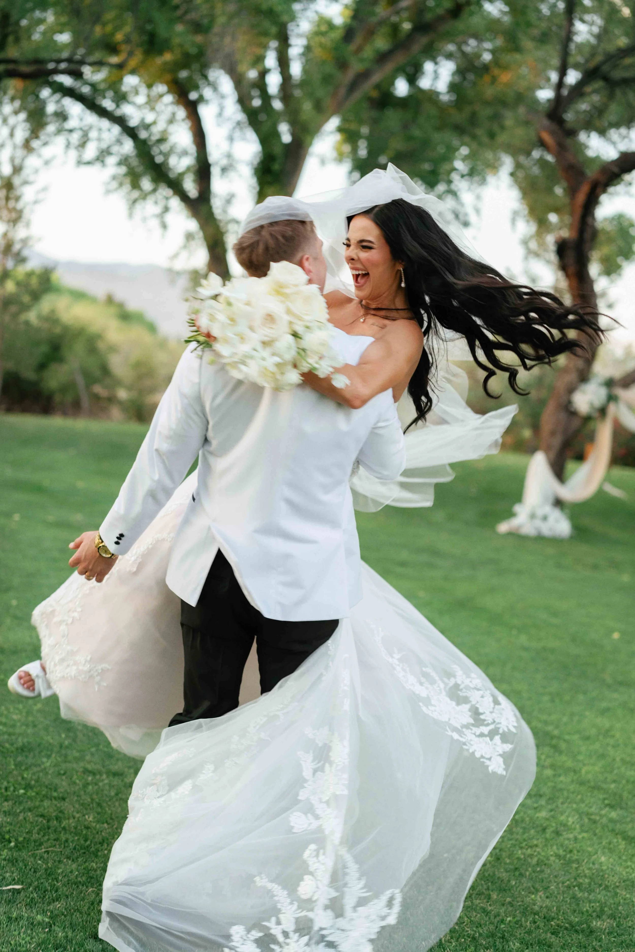 A bride and groom are dancing outdoors on a green lawn with trees in the background. The bride has long black hair, is wearing a strapless white wedding gown with lace details, and is smiling joyfully. The groom is holding the bride and is dressed in