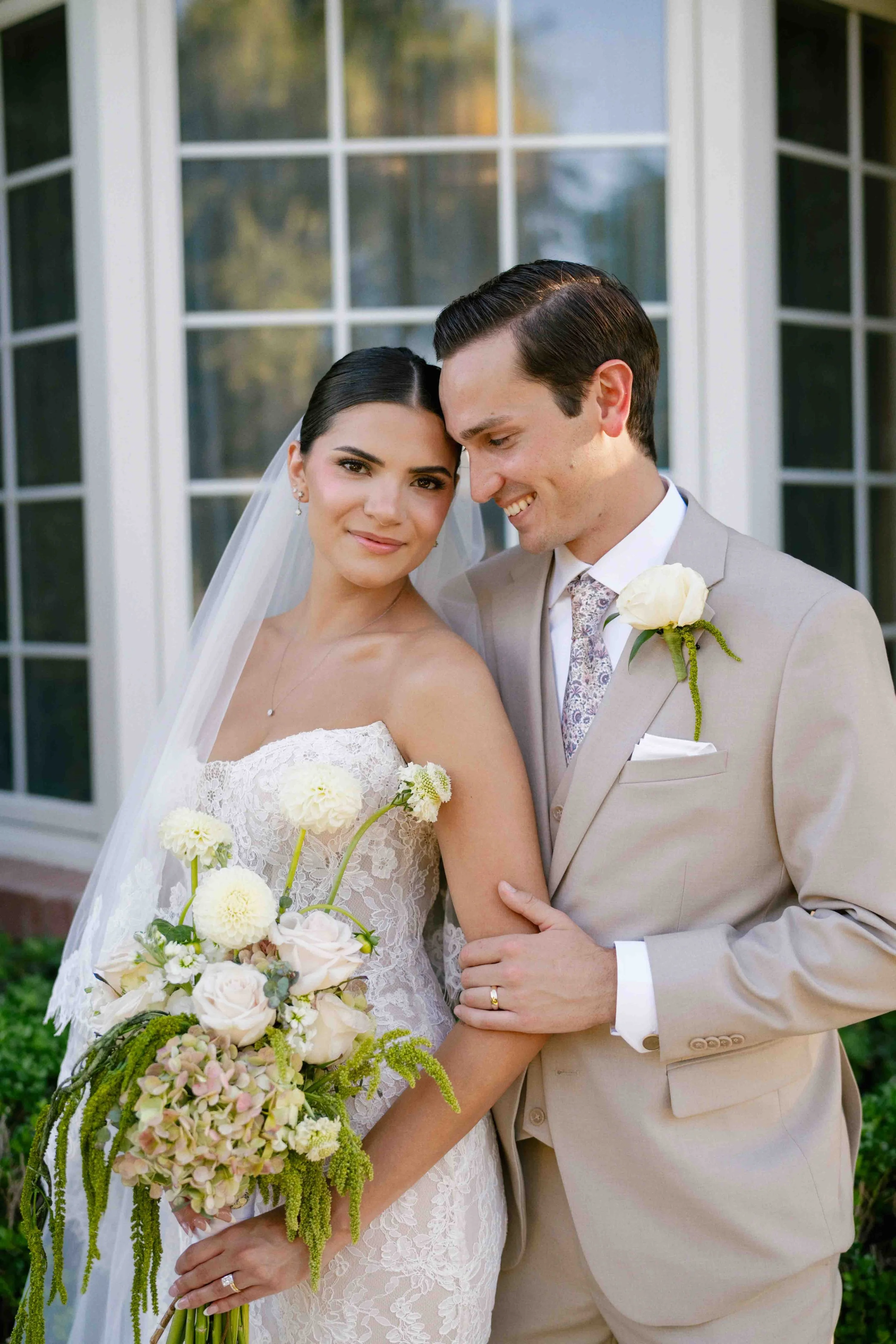 A bride and groom on their wedding day standing close together, holding a floral bouquet, smiling, outdoors in front of a large windowed doorway.