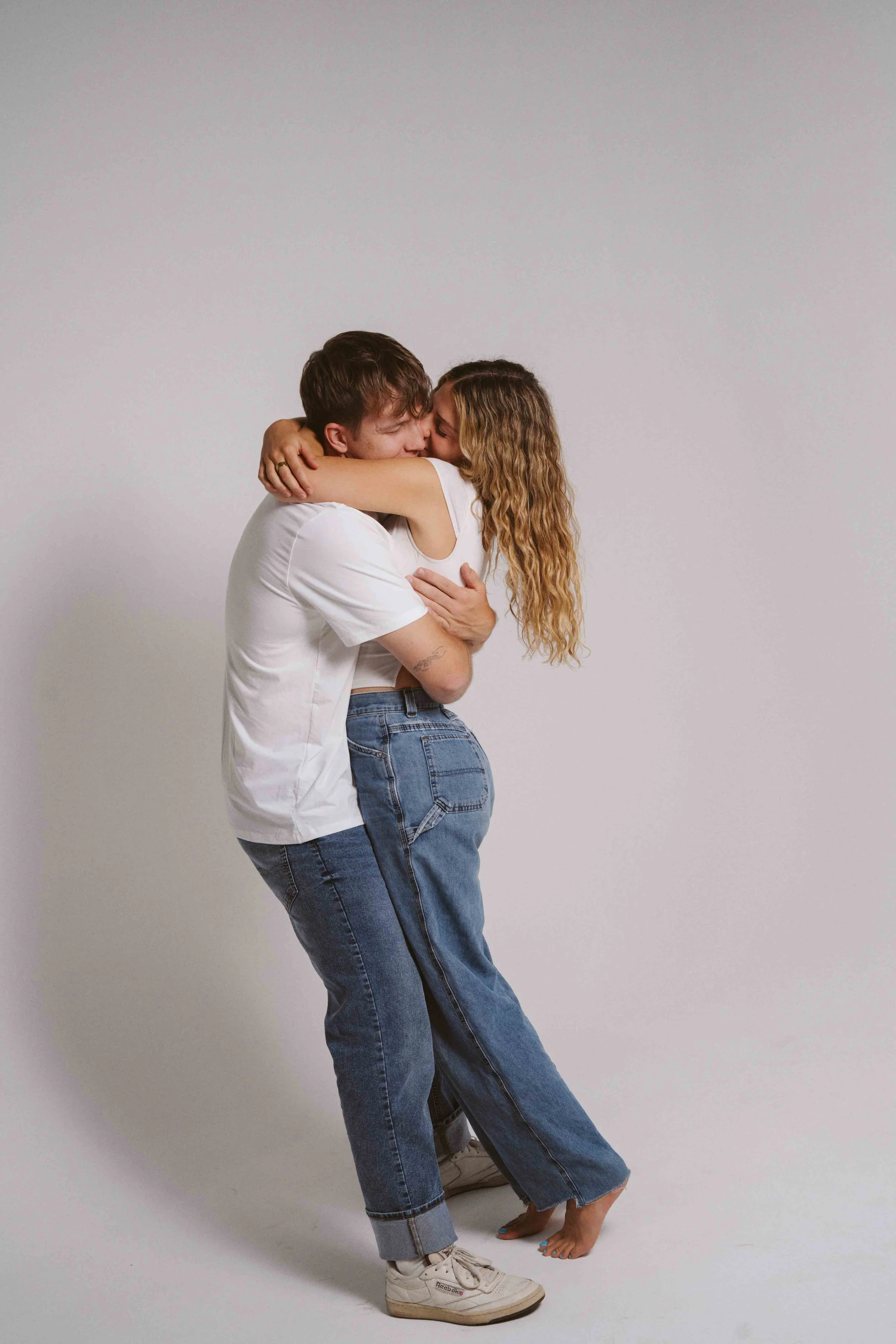 A young man and a young woman are embracing each other in a playful hug against a plain background. The man is wearing a white t-shirt, jeans, and white sneakers. The woman is wearing a white sleeveless top and jeans, with barefoot on one foot and th