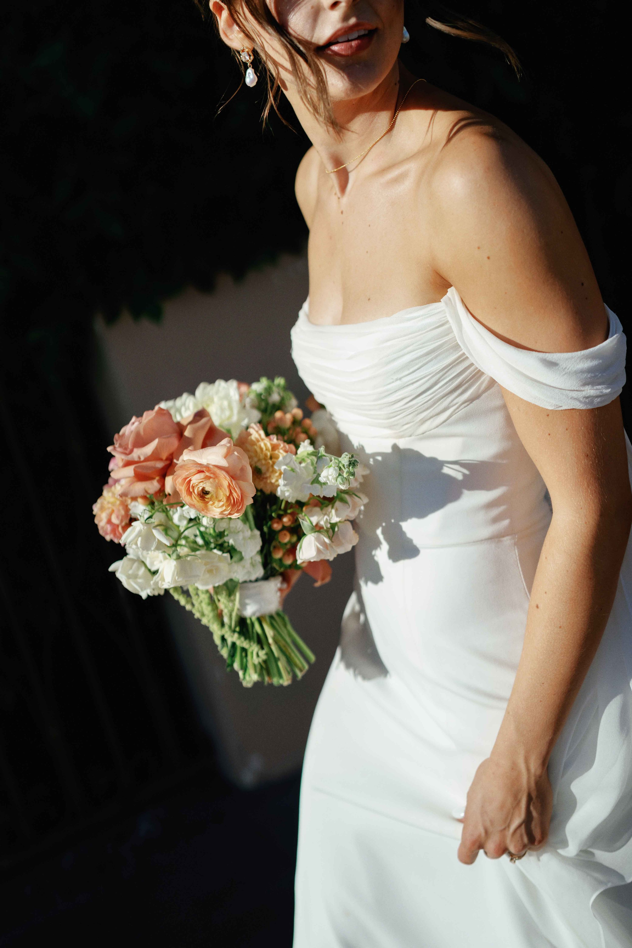 A woman in a white off-shoulder dress holding a bouquet of flowers, with her face partially visible, showing lips and earrings, in a close-up shot.