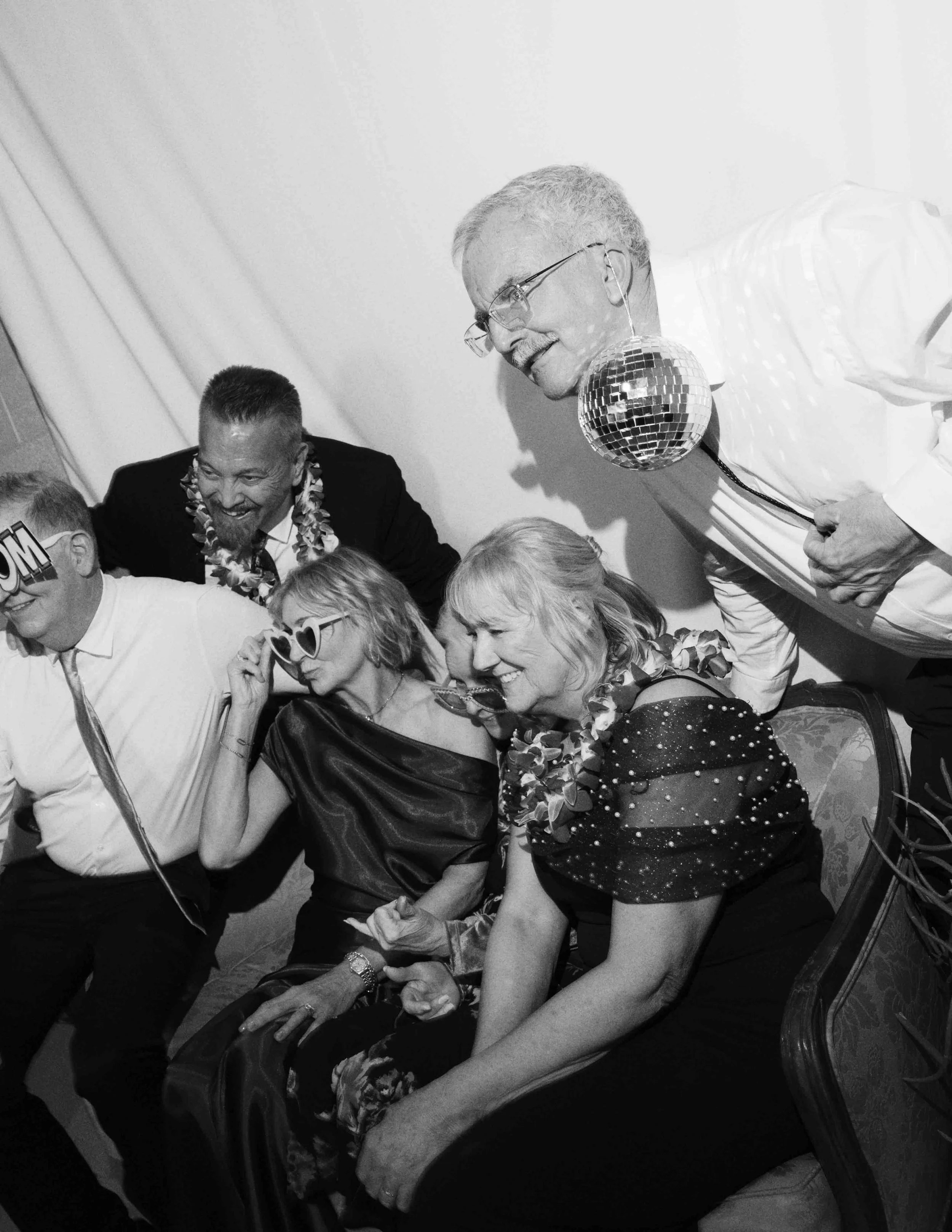 A group of people at a party, wearing festive attire and accessories, with some dancing and smiling. One man is wearing a disco ball necklace, and a woman is adjusting her glasses. The scene has a lively, cheerful atmosphere.