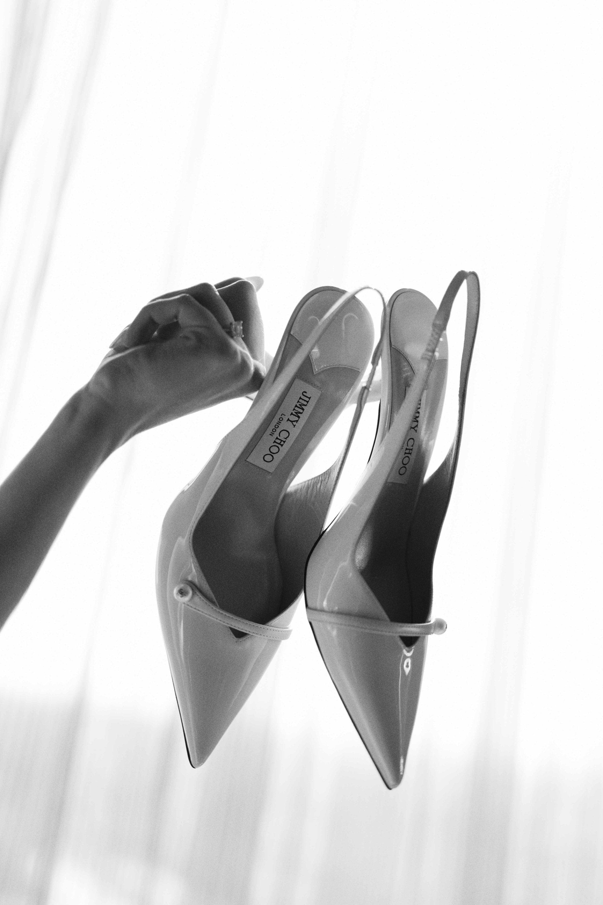 Black and white photo of a hand holding mismatched white high-heeled shoes from Jimmy Choo, hanging against a light background.