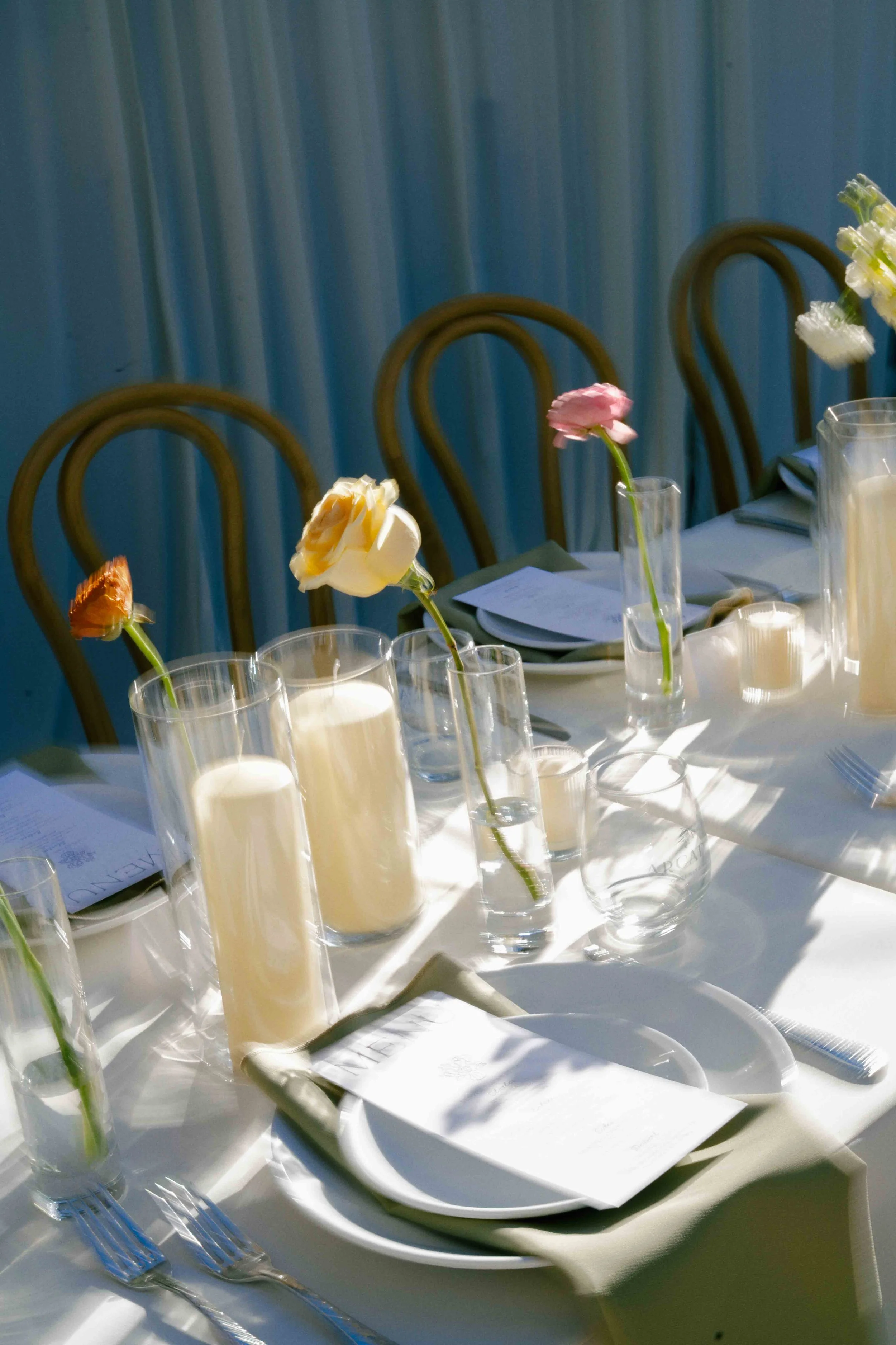 A table decorated with flowers in tall glass vases, surrounded by white plates, silverware, and candles, with a blue curtain backdrop.