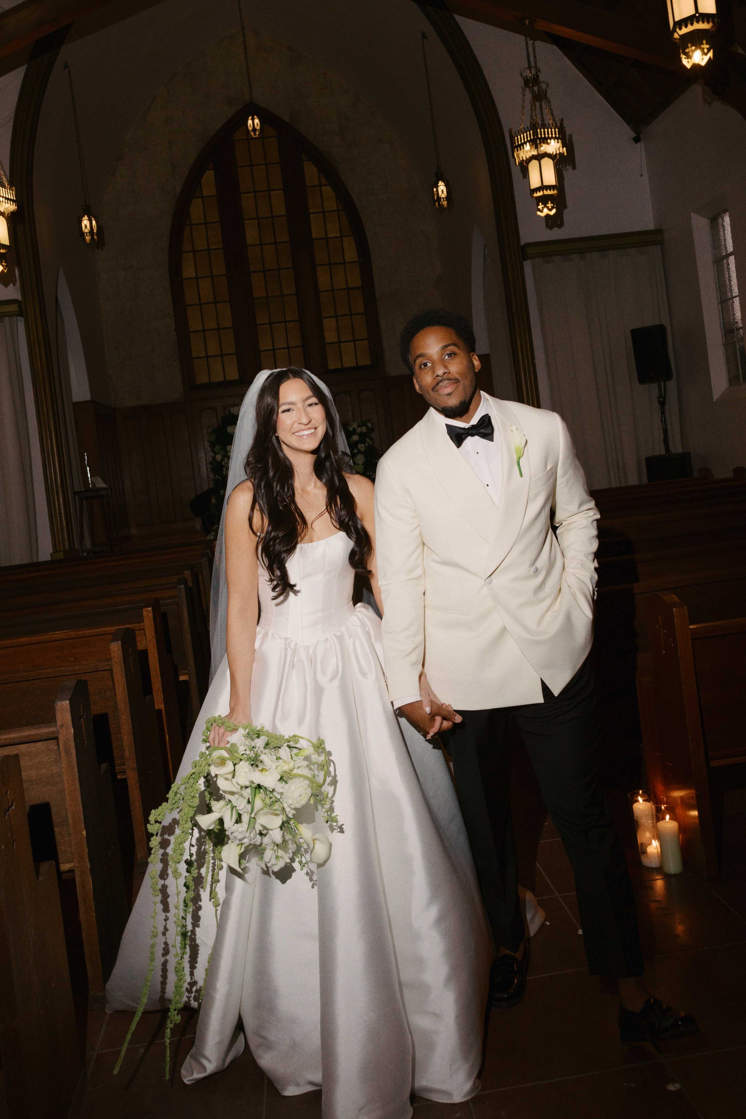 A bride and groom standing inside a church, smiling and holding hands. The bride is wearing a white wedding dress and holding a large bouquet of white flowers, and the groom is dressed in a white tuxedo with a black bow tie. There are candles on the 