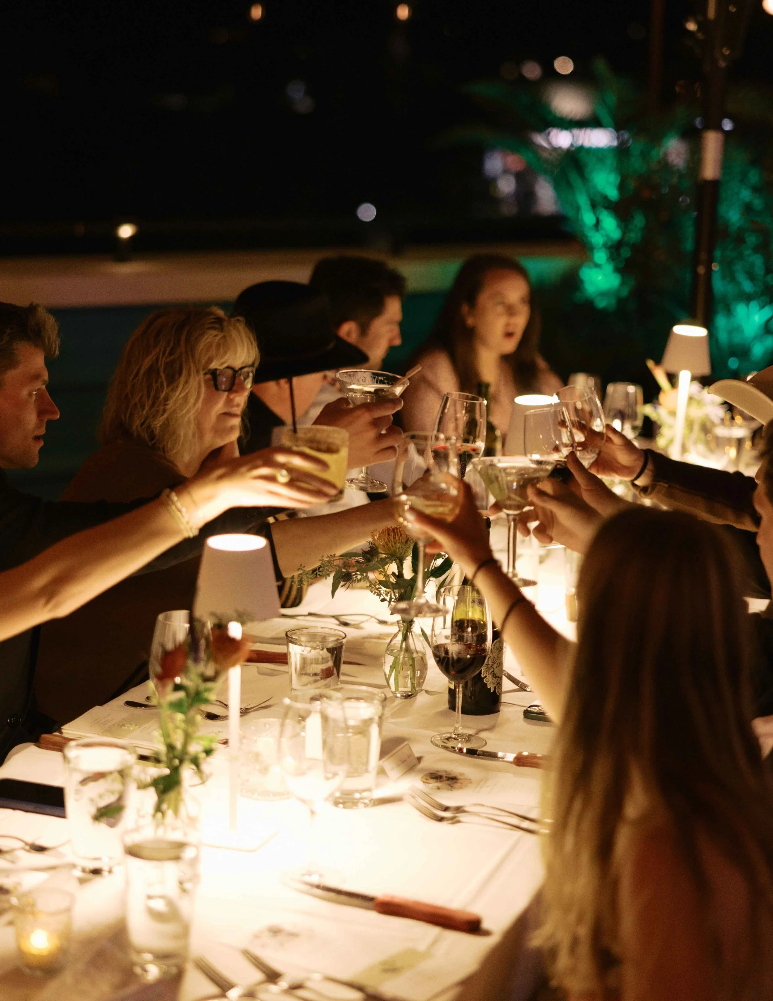 People sitting at a formal dinner table outdoors at night, raising glasses for a toast. The table is decorated with flowers and lit by candlelight, with palm trees and lights visible in the background.