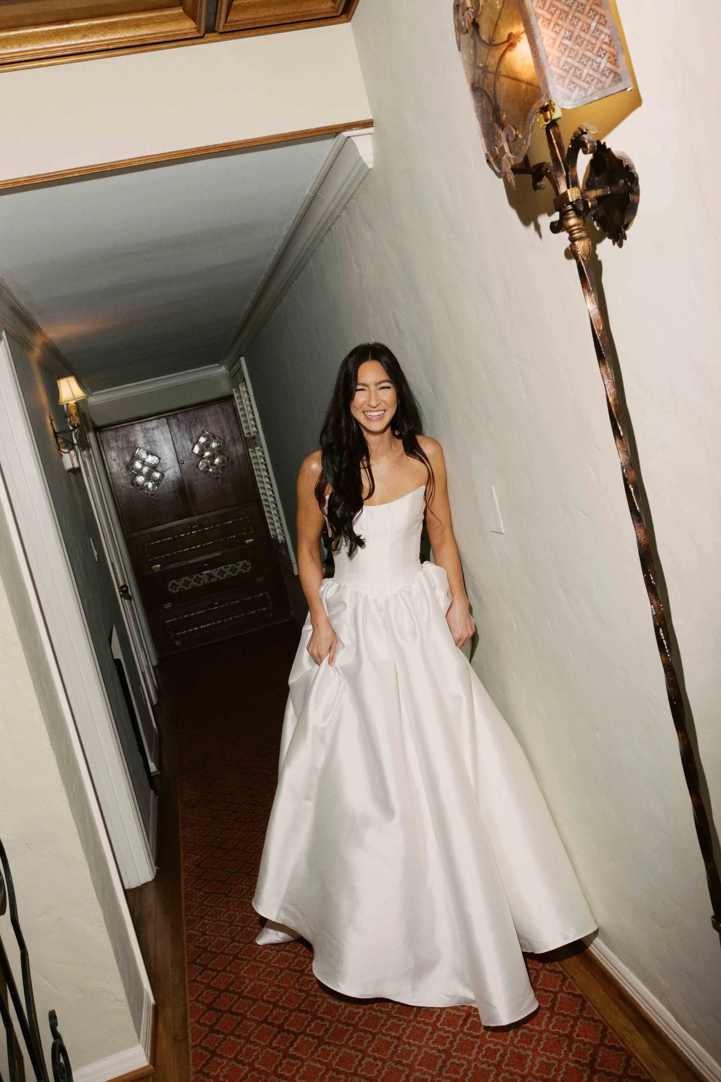 A woman in a white strapless wedding dress standing in a hallway, smiling and holding the sides of her dress.