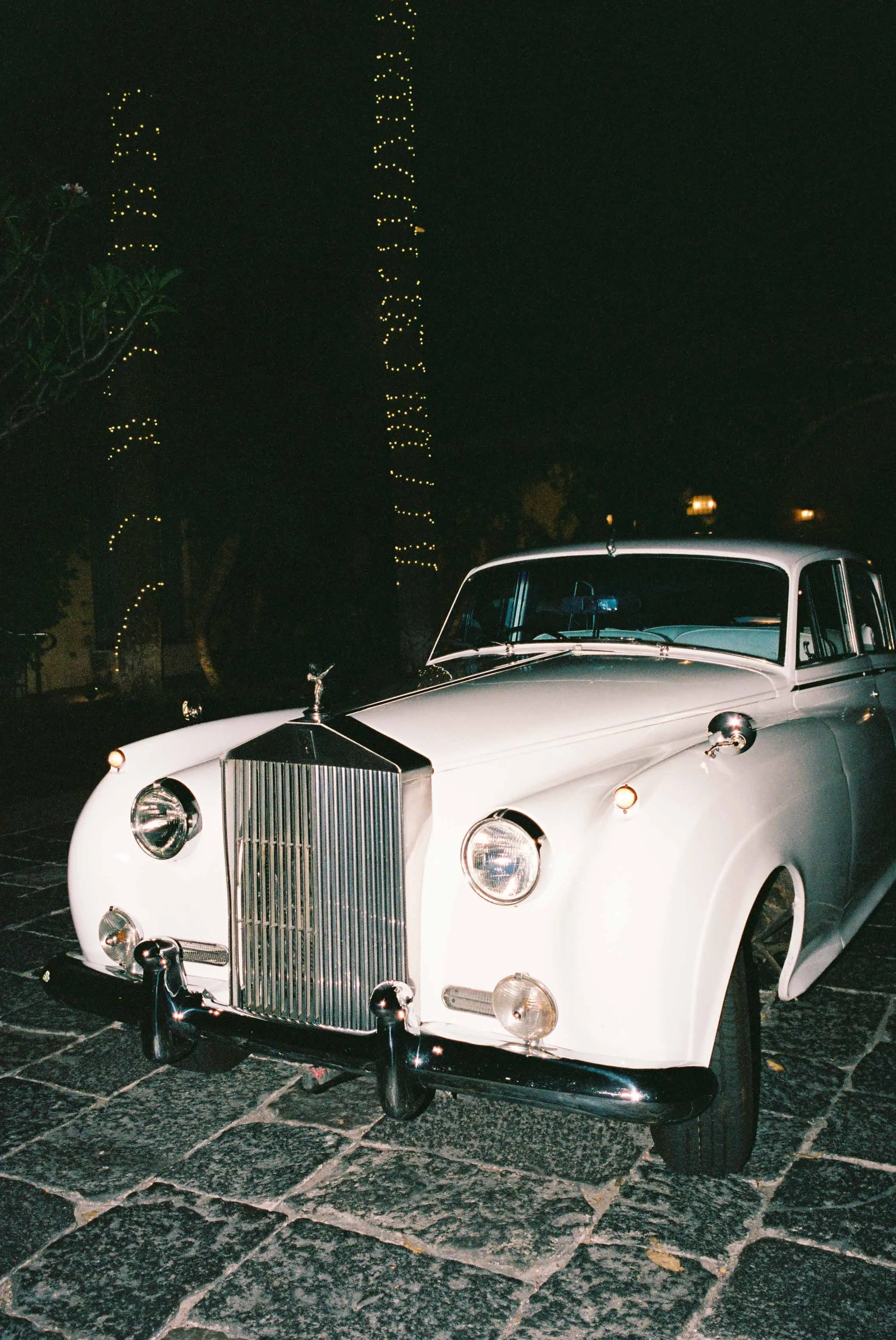 Vintage white luxury car parked on a stone-paved street at night with palm trees decorated in string lights in the background.