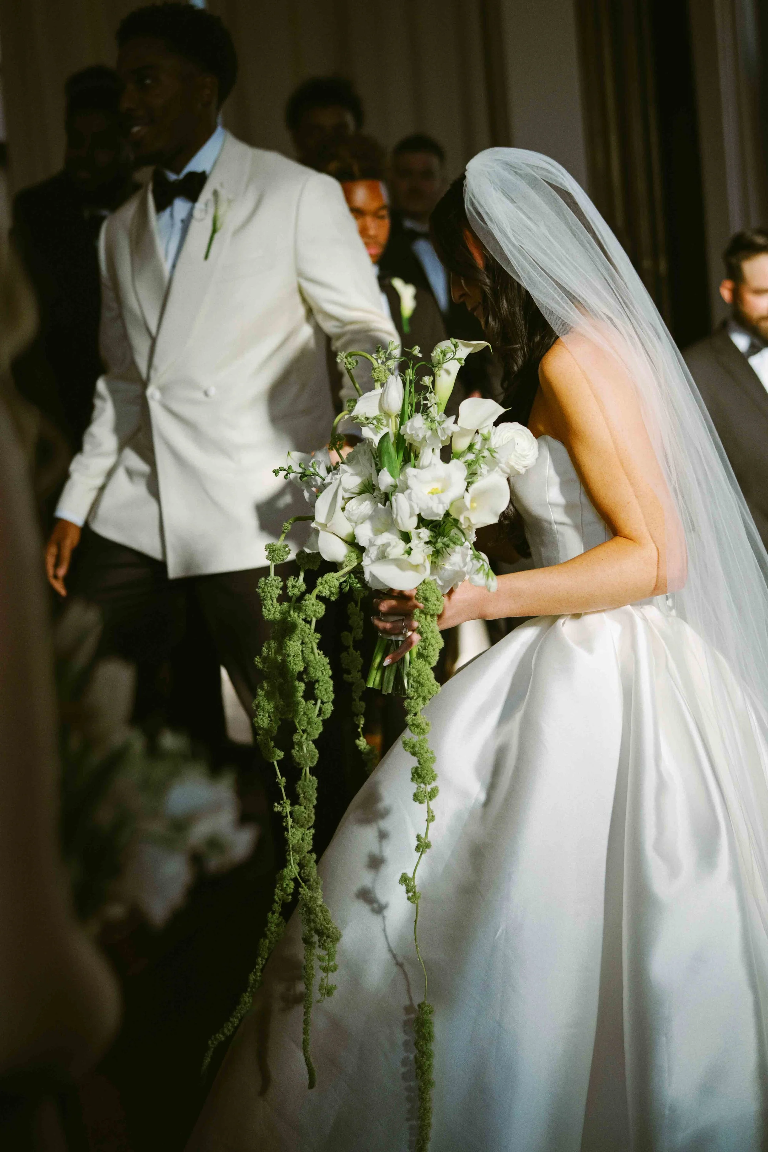 A bride in a white wedding gown and veil holding a bouquet of white flowers, standing beside a groom dressed in a white tuxedo with a black bow tie, during a wedding ceremony.