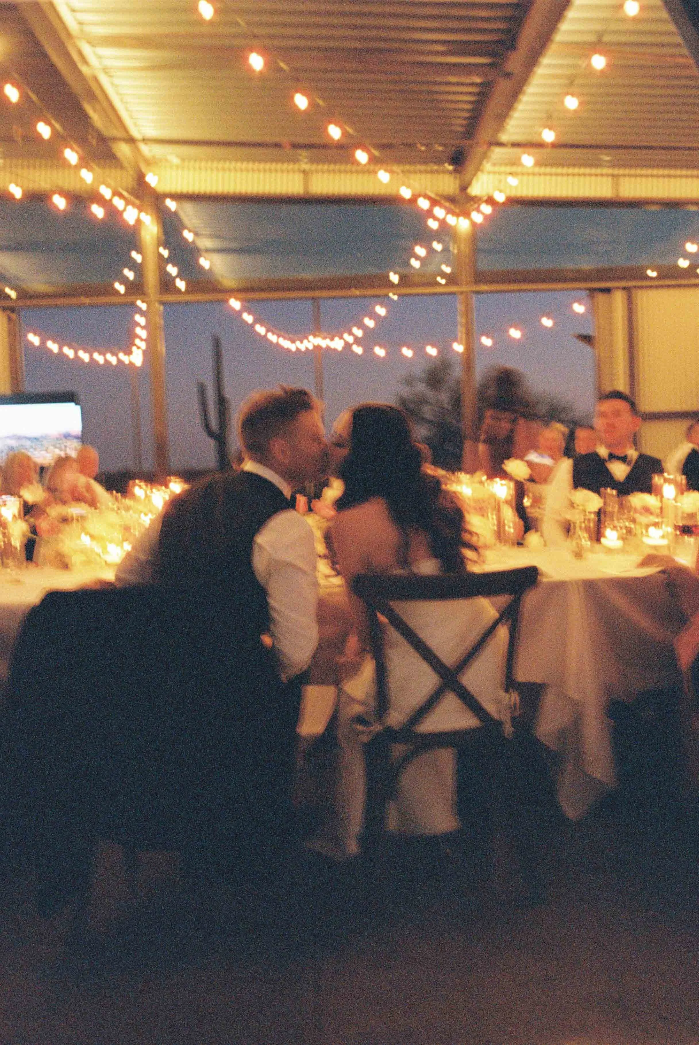 A group of people in formal attire dining at a table illuminated by string lights in a semi-open space during evening or night.