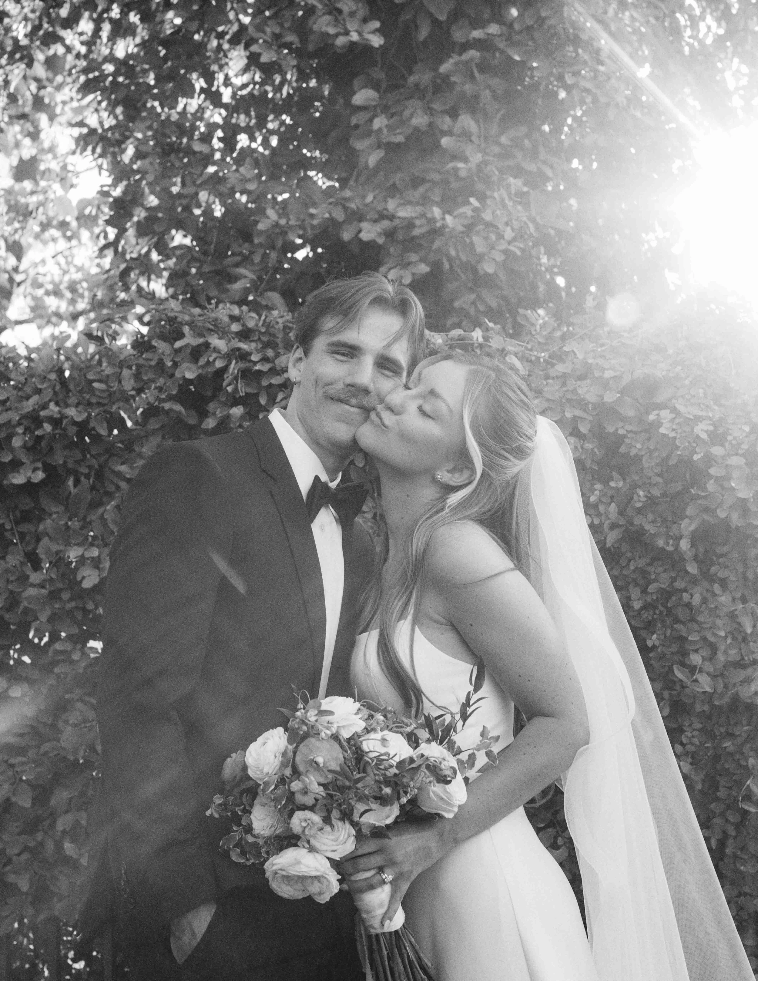 A black and white photo of a newly married couple standing outdoors in front of bushes, with the man in a tuxedo and the woman in a wedding dress holding a bouquet of flowers, kissing each other.