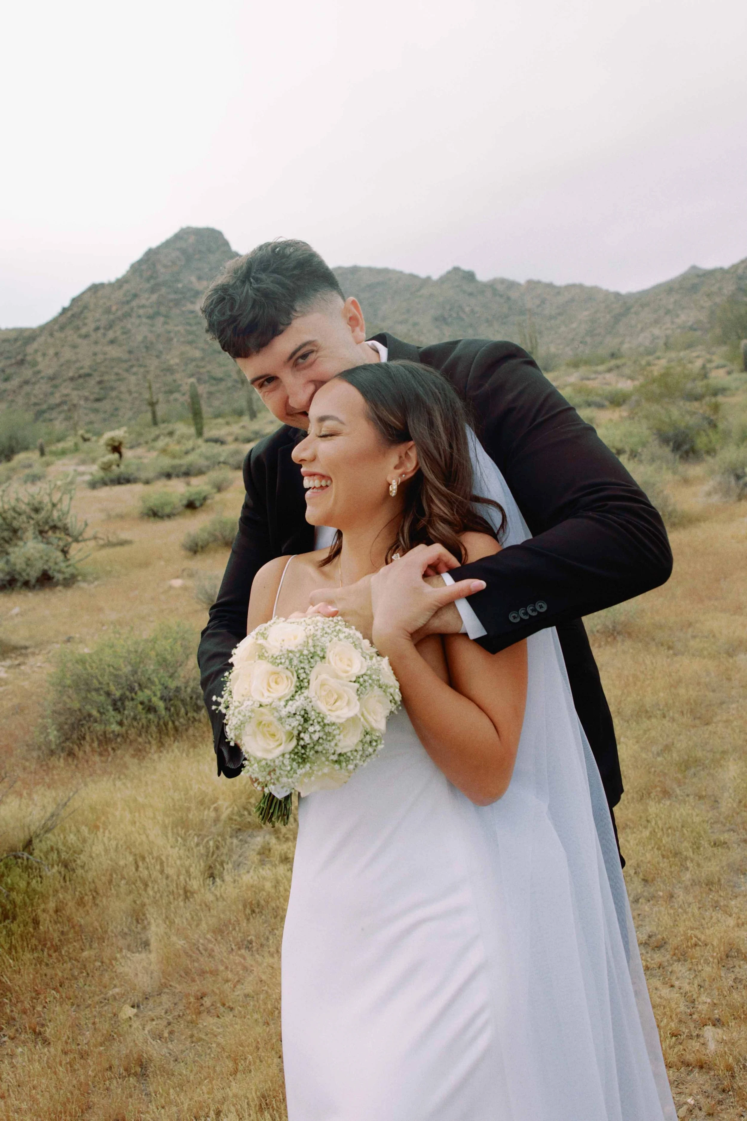 A bride and groom embrace in a desert landscape with cacti and mountains in the background, both smiling and happy.