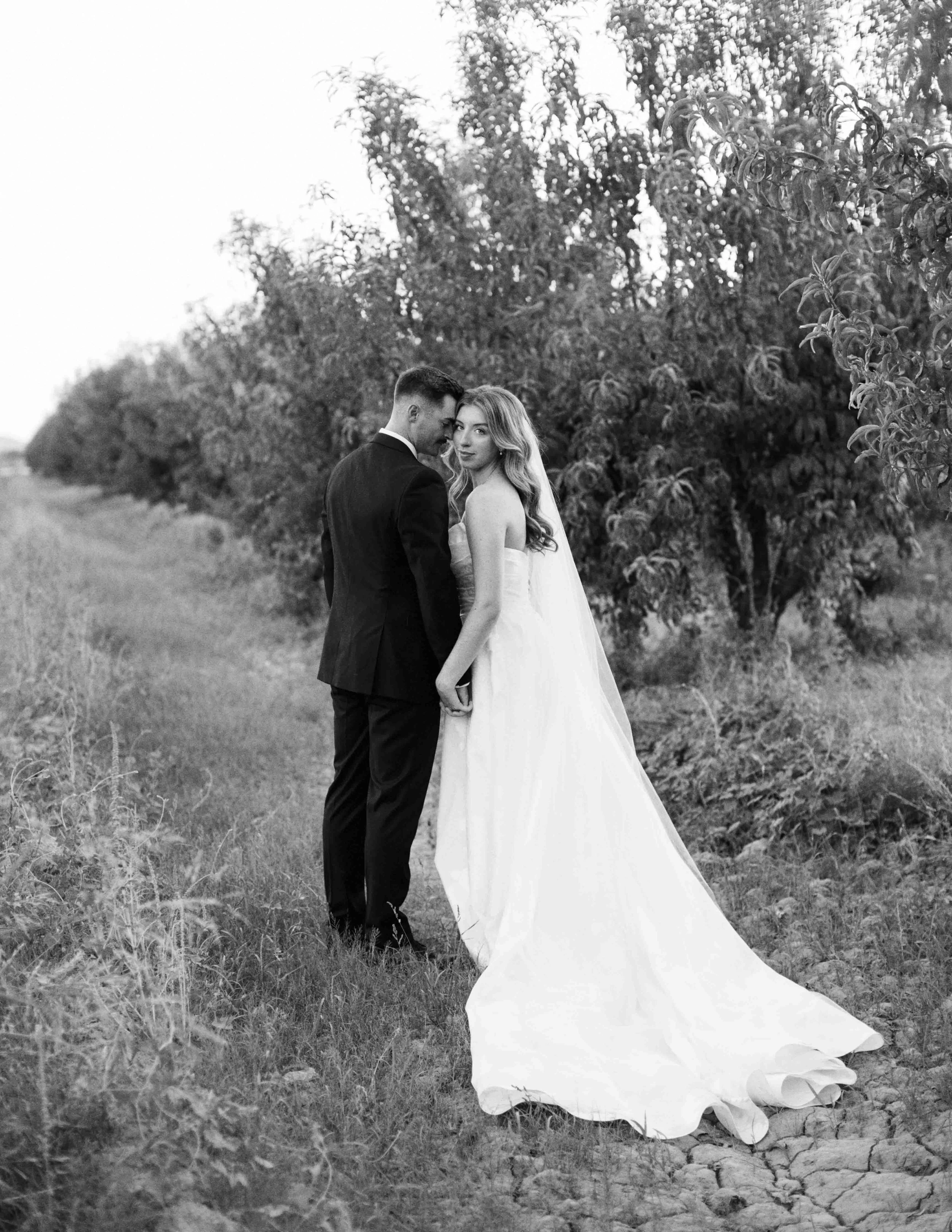 A black and white photograph of a bride and groom standing outdoors, holding hands, with the groom leaning in to whisper to the bride, who looks at the camera. There are trees and bushes in the background, and the bride's dress has a long train on th