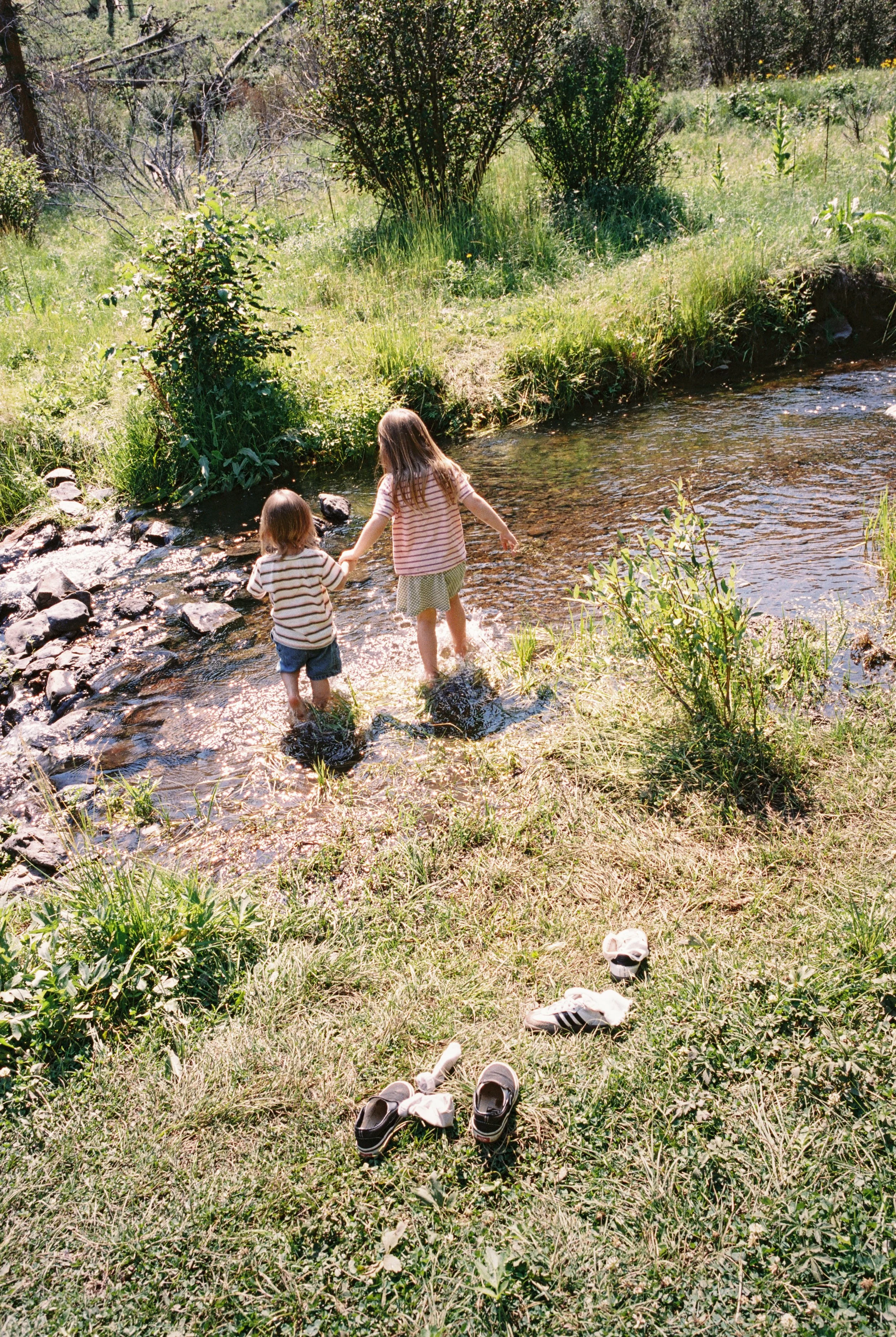 Two children wading in a creek in a grassy, wooded area with shoes scattered on the grass nearby.