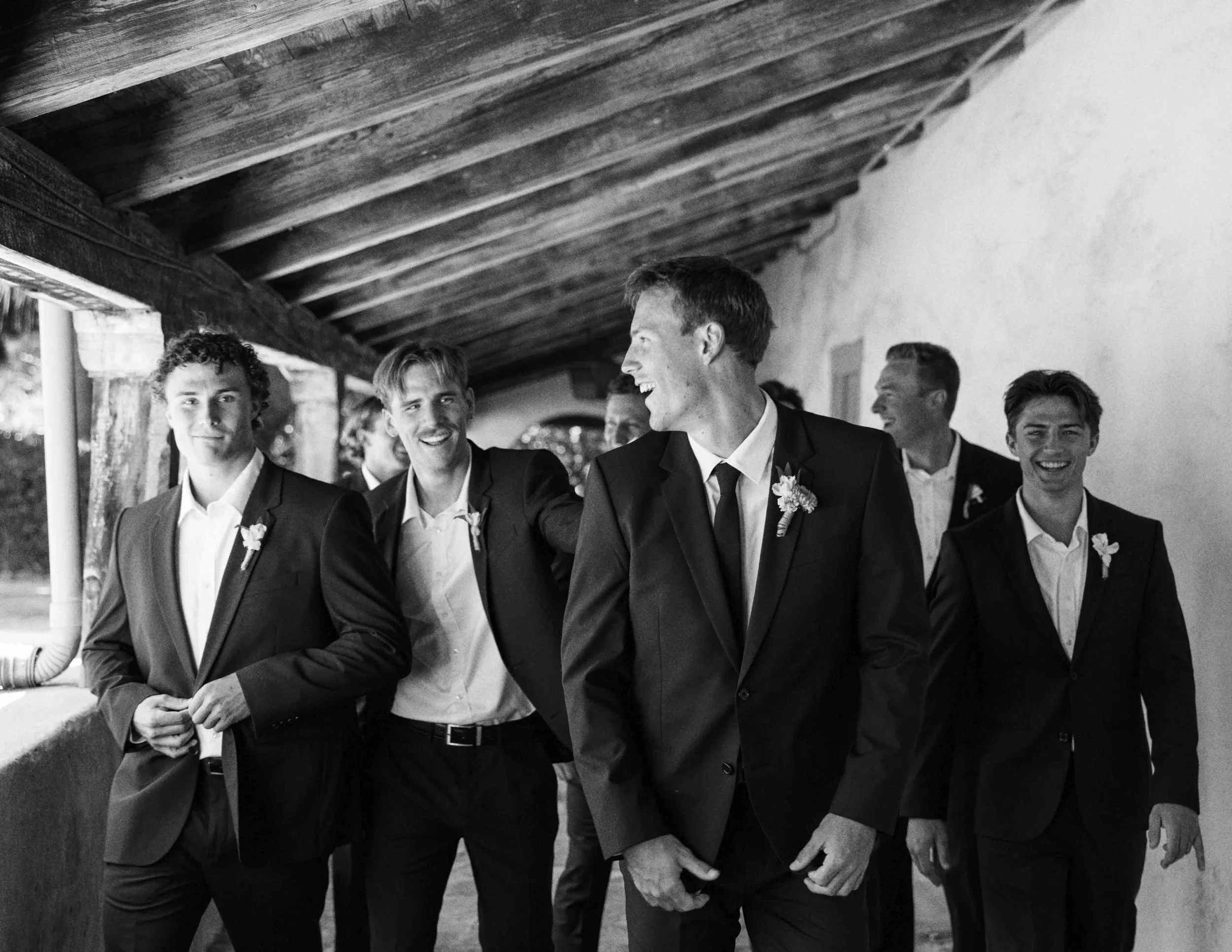 A group of men dressed in suits, some with bow ties, standing under a wooden structure, engaged in conversation and smiling.