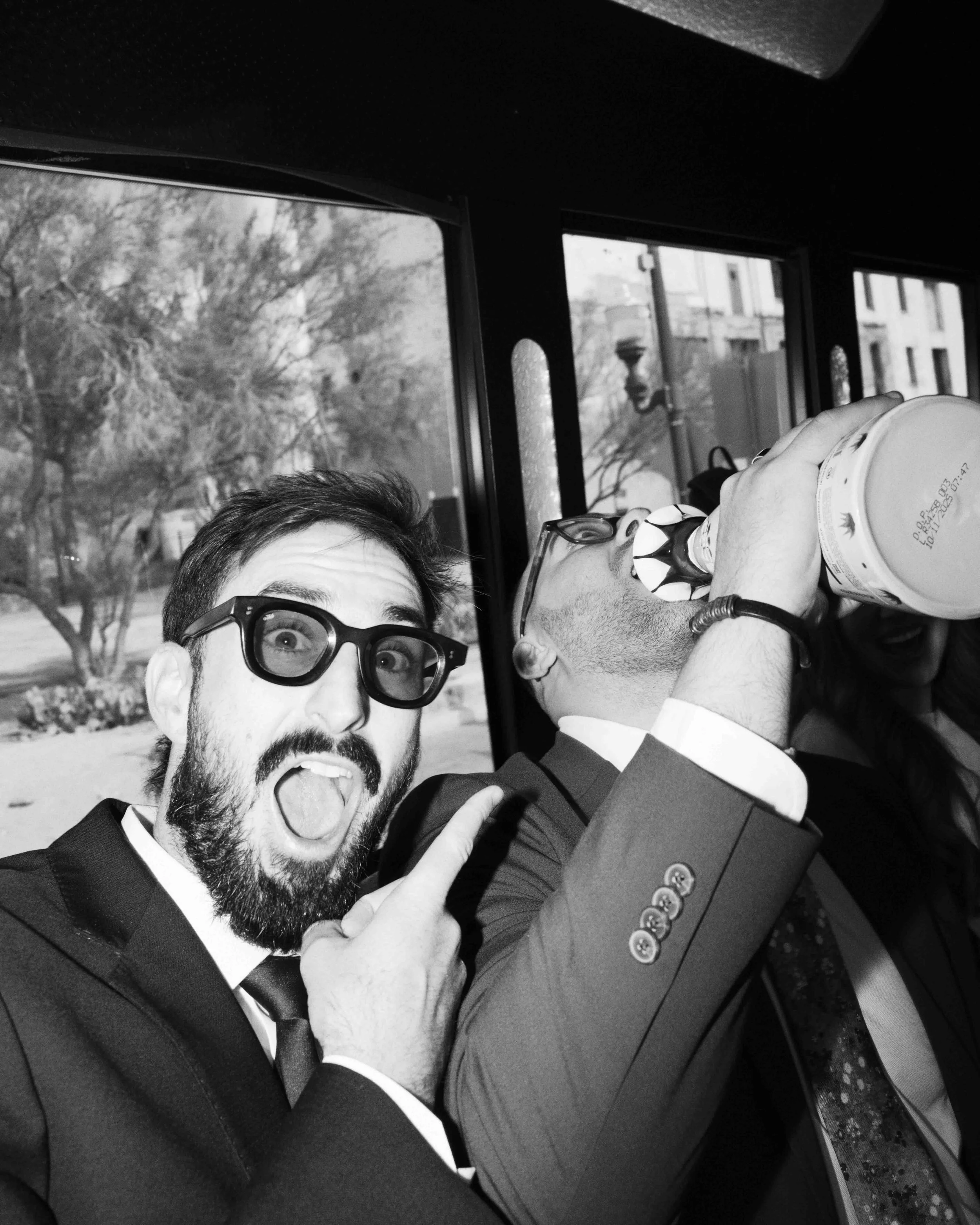 Two men in suits and glasses appear to be celebrating with a toast, holding a drink close to their mouths inside a bus or train.