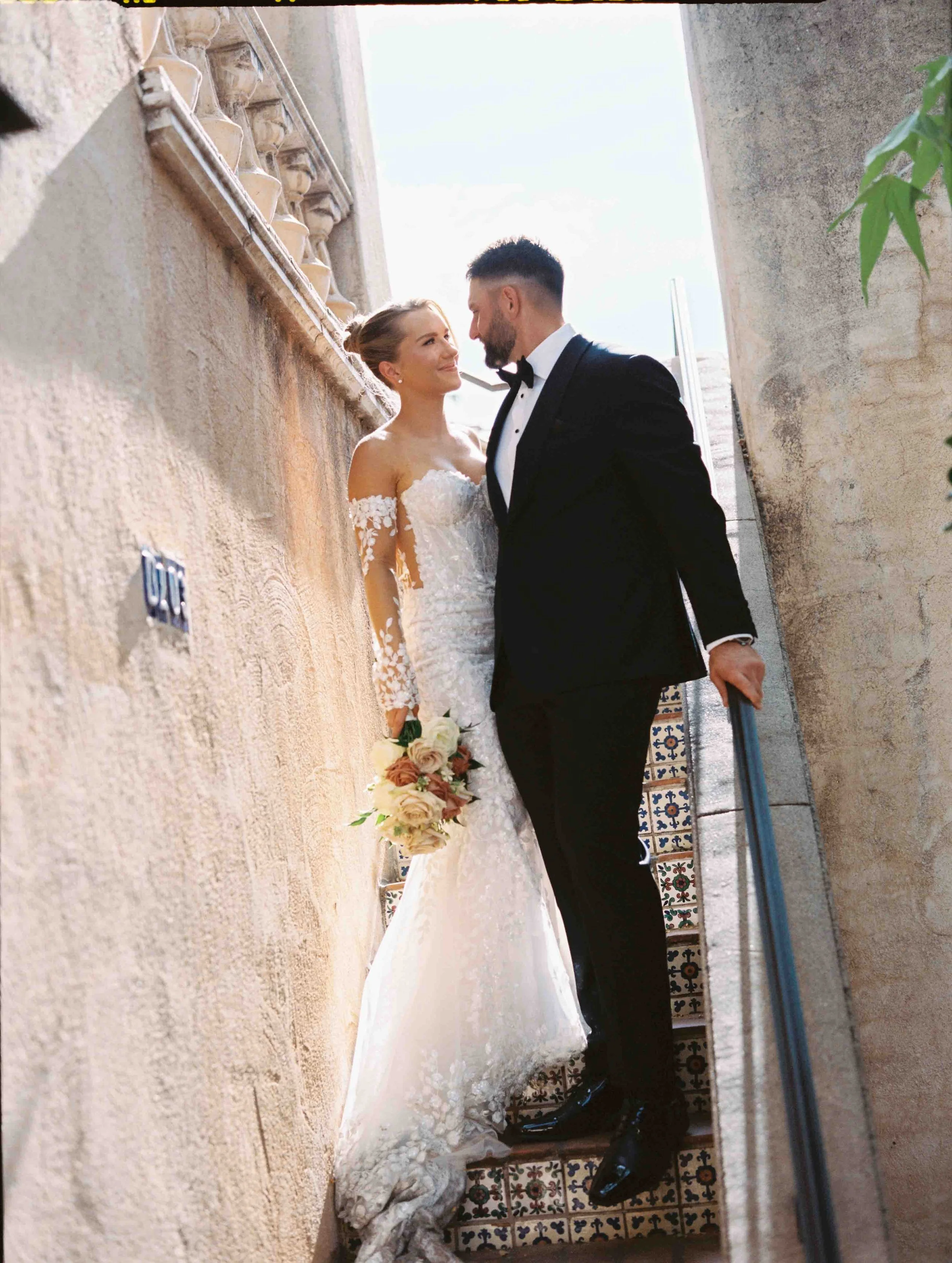 A bride and groom in wedding attire are standing close together in an outdoor corridor with textured walls and decorative tiled flooring. The bride is holding a bouquet of flowers and looking lovingly at the groom, who is wearing a tuxedo. They are s