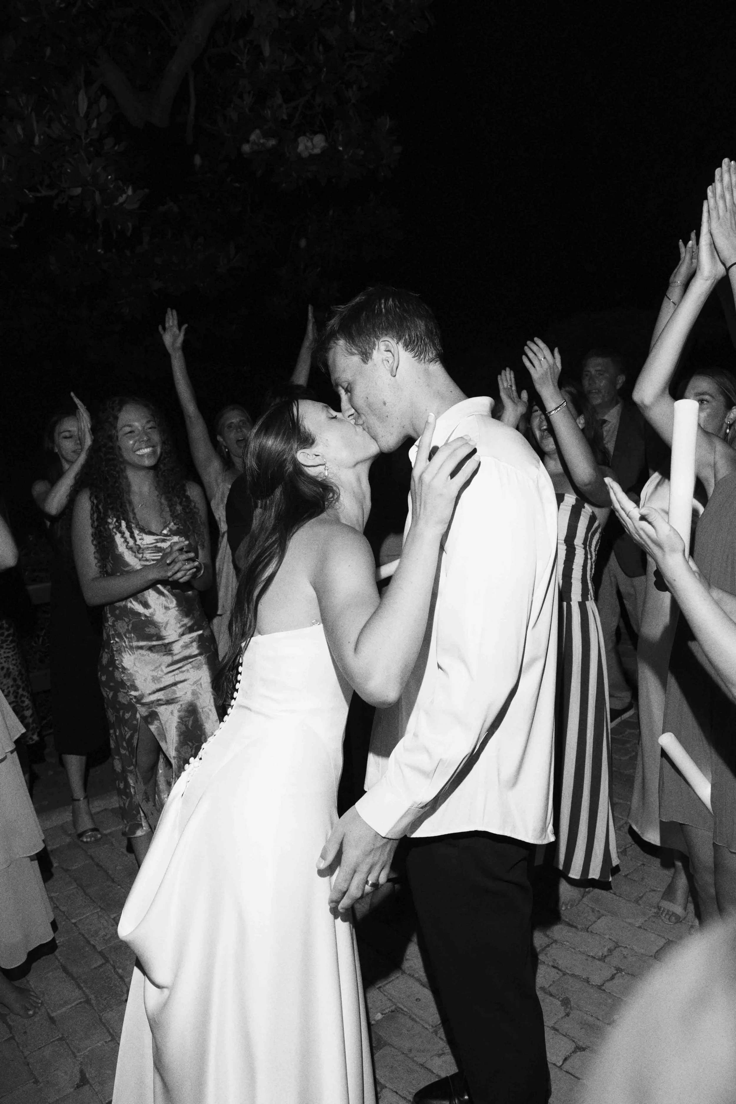 A black and white photo of a bride and groom sharing a kiss at their wedding reception, surrounded by friends celebrating and dancing.