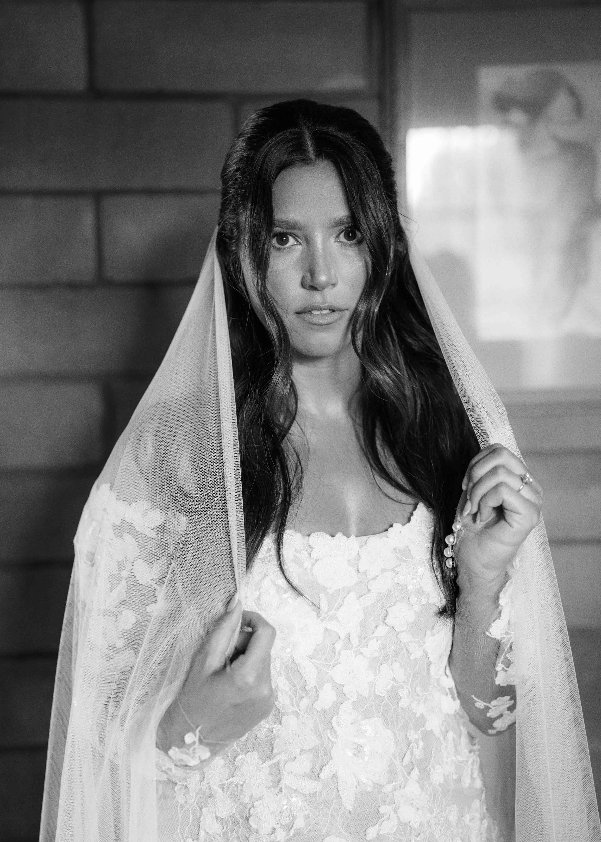 Black and white photograph of a woman in a wedding dress with lace details, wearing a veil, standing indoors. She has long dark hair and is holding a rosary in her right hand, looking directly at the camera.