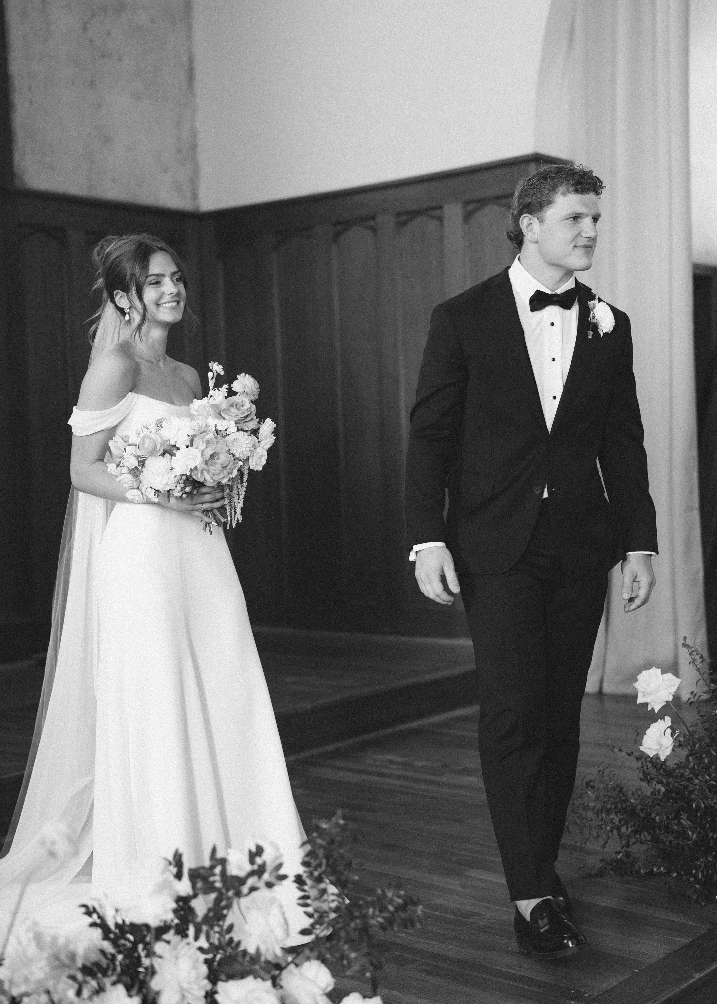 Black and white photo of a bride and groom on their wedding day. The bride is smiling, holding a bouquet, and wearing an off-shoulder wedding dress with a veil. The groom is walking ahead, wearing a tuxedo with a bow tie and boutonniere.