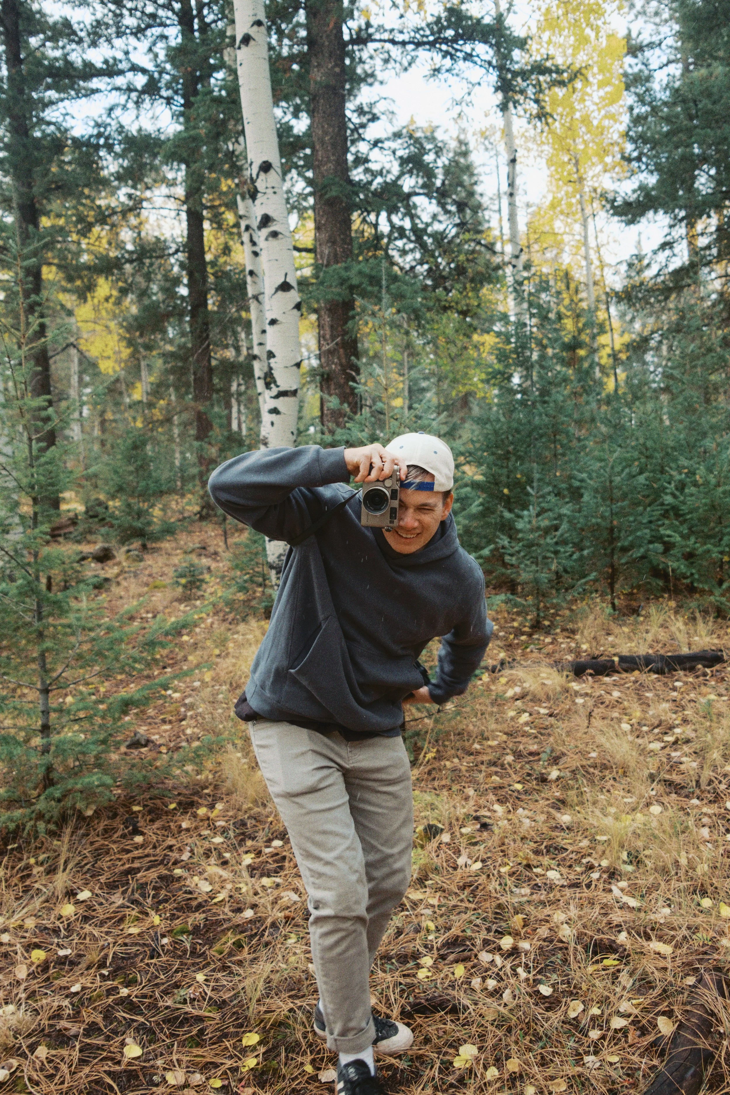 A person with a backwards white cap taking a photo with a camera in a forest with tall trees, some with white bark, and a ground covered with fallen leaves and pine needles.