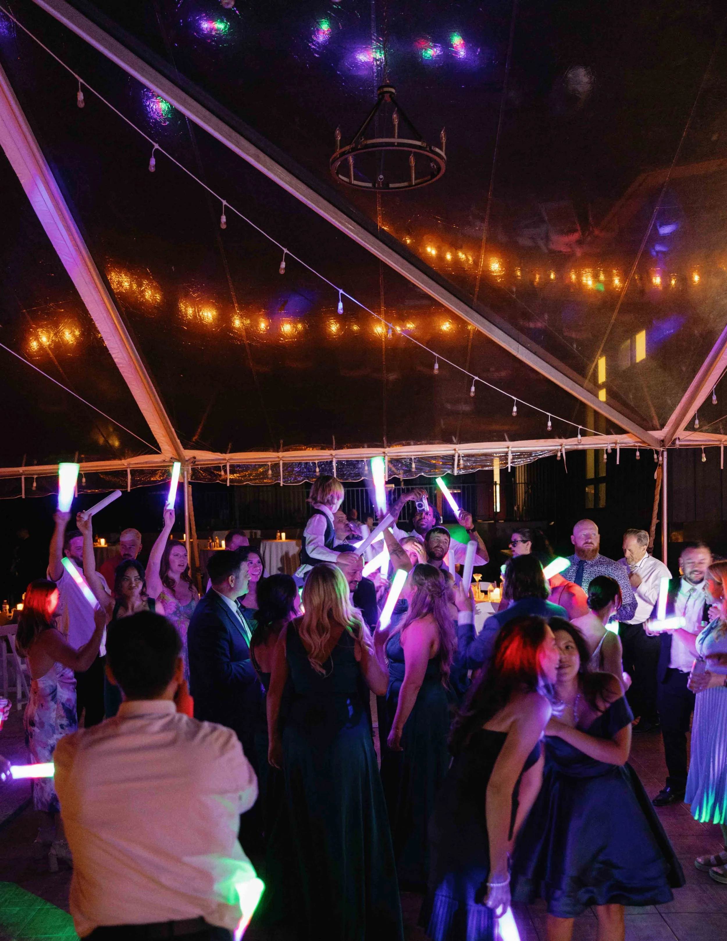 A group of people dancing at a party or celebration under a tent with colorful lights and a chandelier.