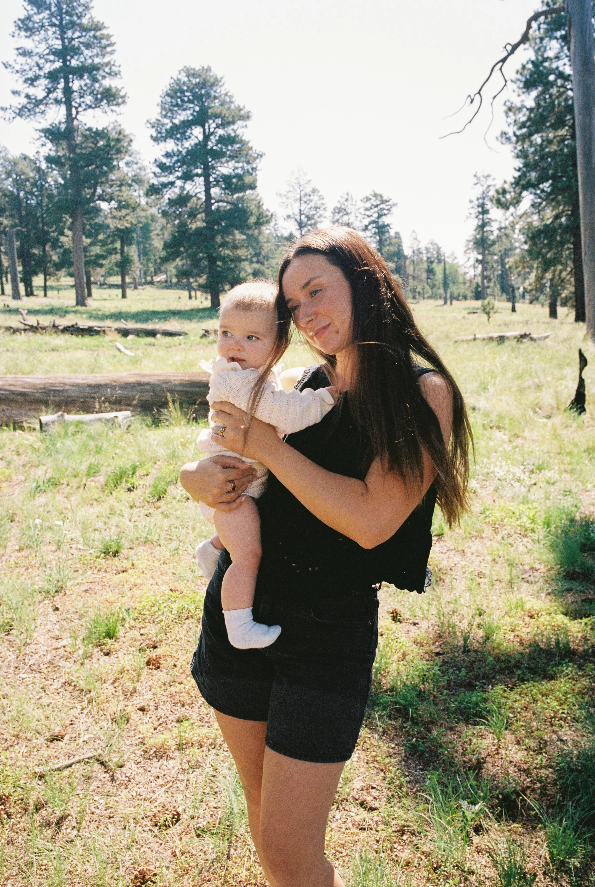 A woman with long brown hair wearing a black sleeveless top and shorts holds a small blonde-haired child with light skin, wearing a cream-colored sweater and white socks, outdoors in a grassy area with trees under a bright sky.