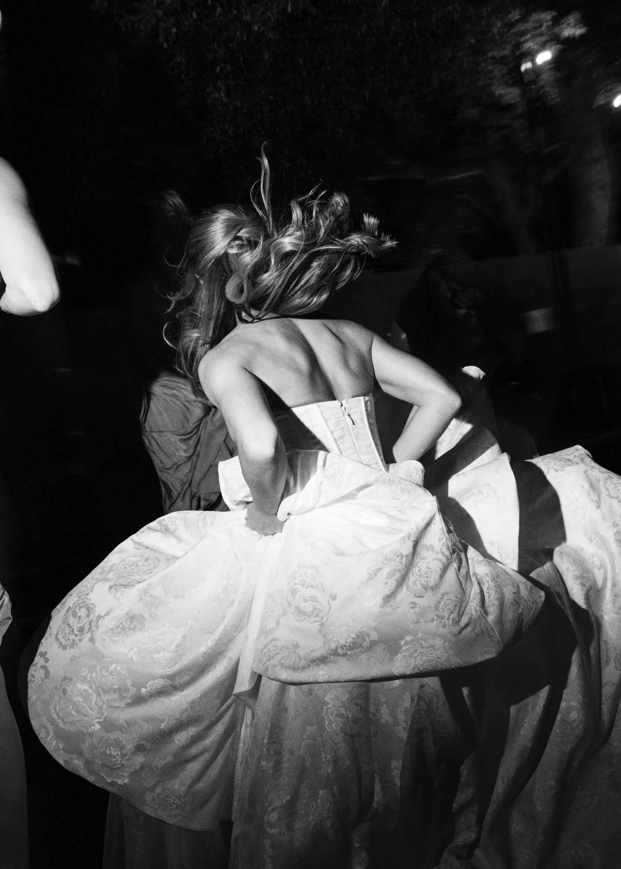 A person with long hair is leaning forward and wearing a strapless dress, sitting on a chair, with their back exposed. They are surrounded by flowing fabric with a floral pattern. The image is black and white, taken in a dark environment.