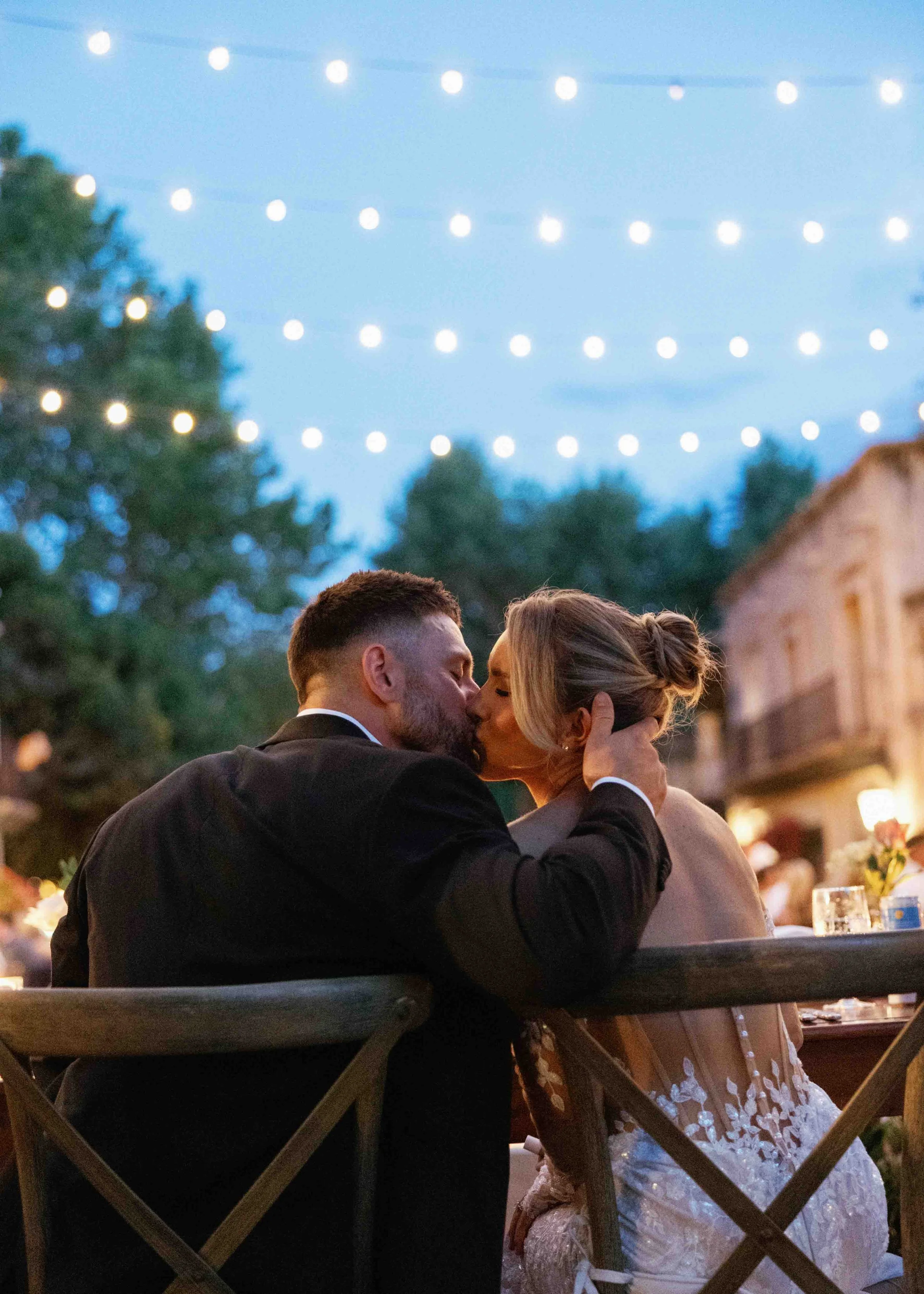 A couple sharing a kiss at an outdoor evening event with string lights overhead and trees in the background.