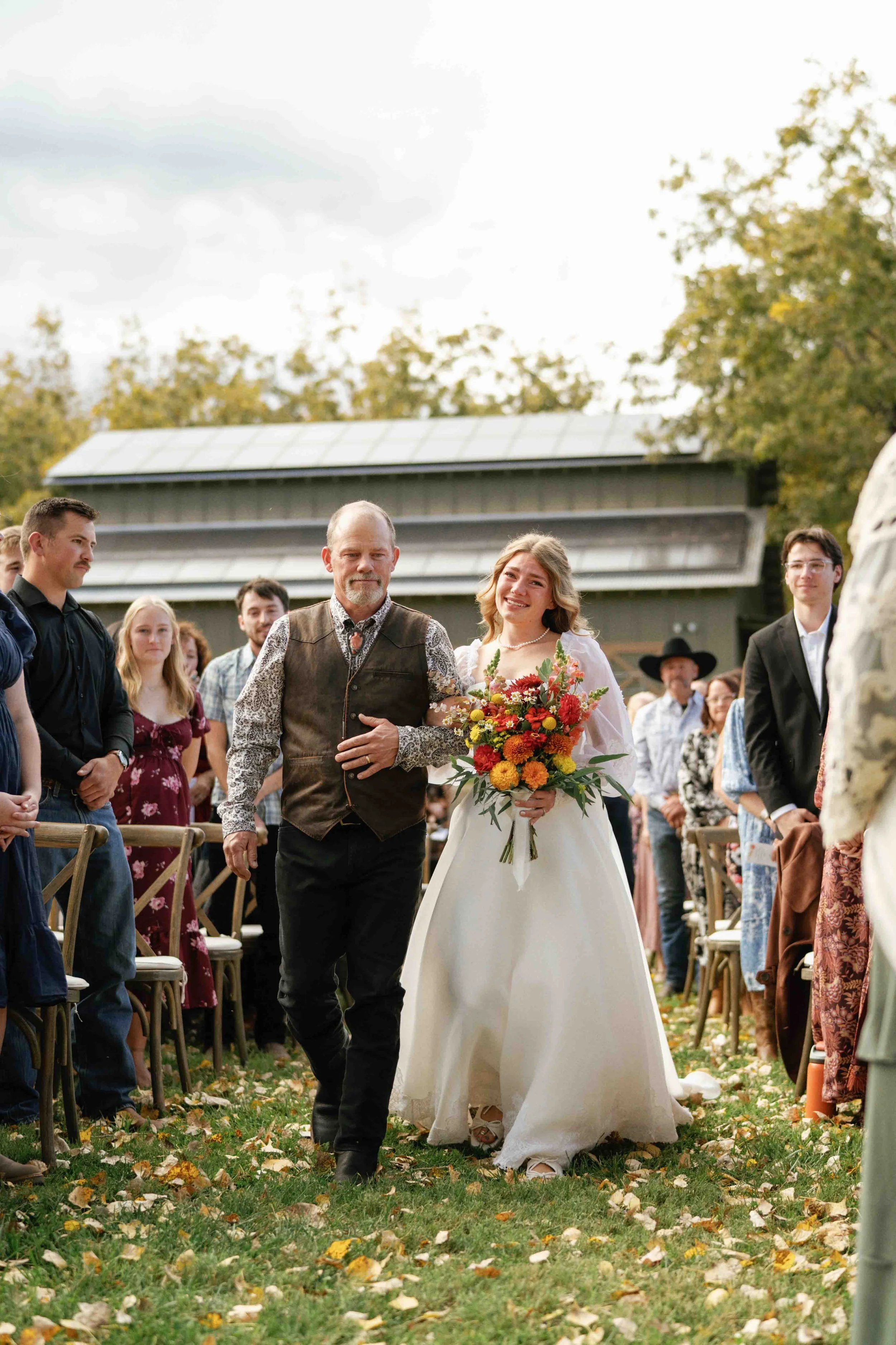 A wedding ceremony outdoors with a bride holding a colorful bouquet walking down an aisle lined with seated guests and family members, surrounded by fallen autumn leaves.