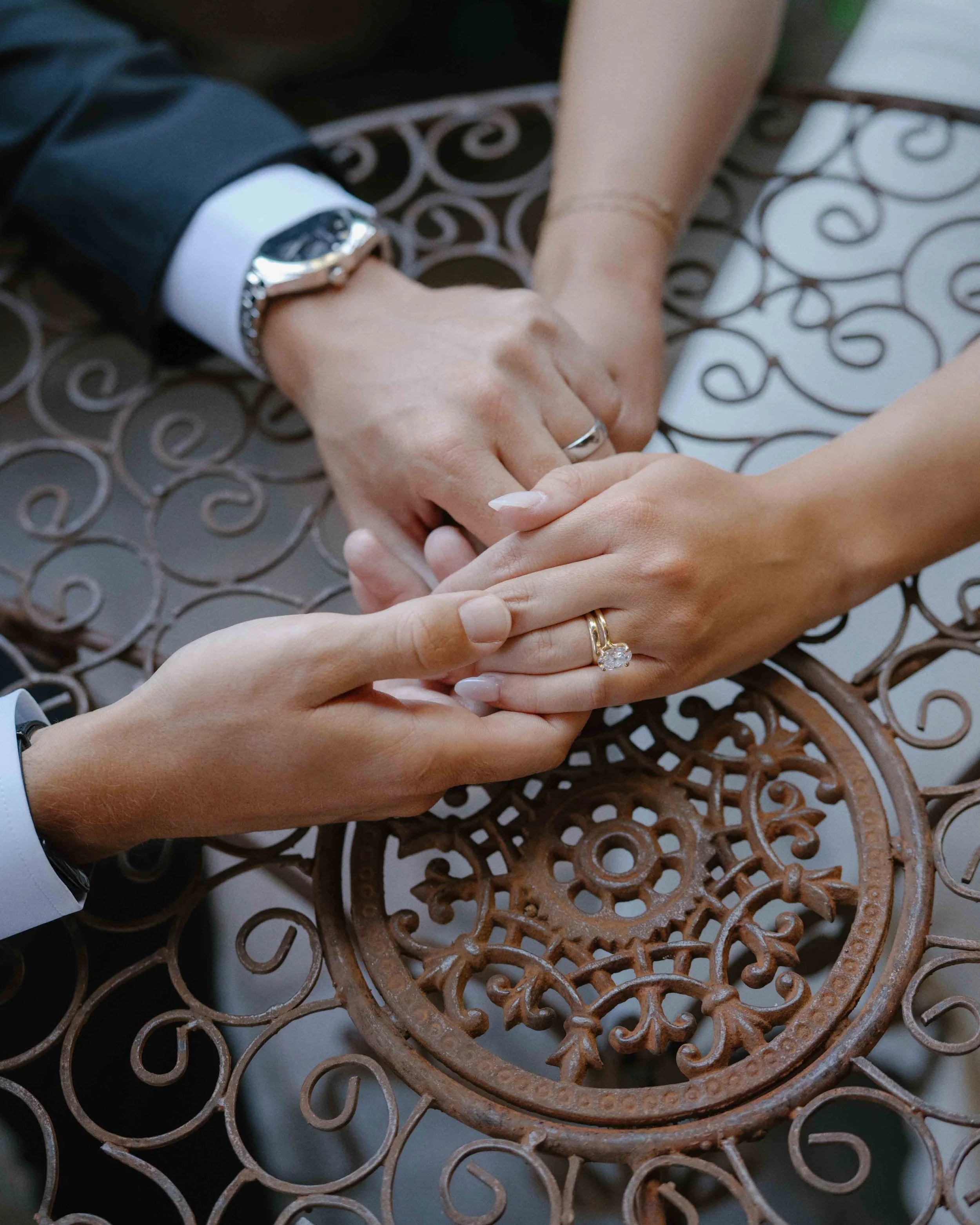 Close-up of two hands, one male and one female, holding each other over a decorative metal table, with the focus on a wedding ring on the woman's finger.