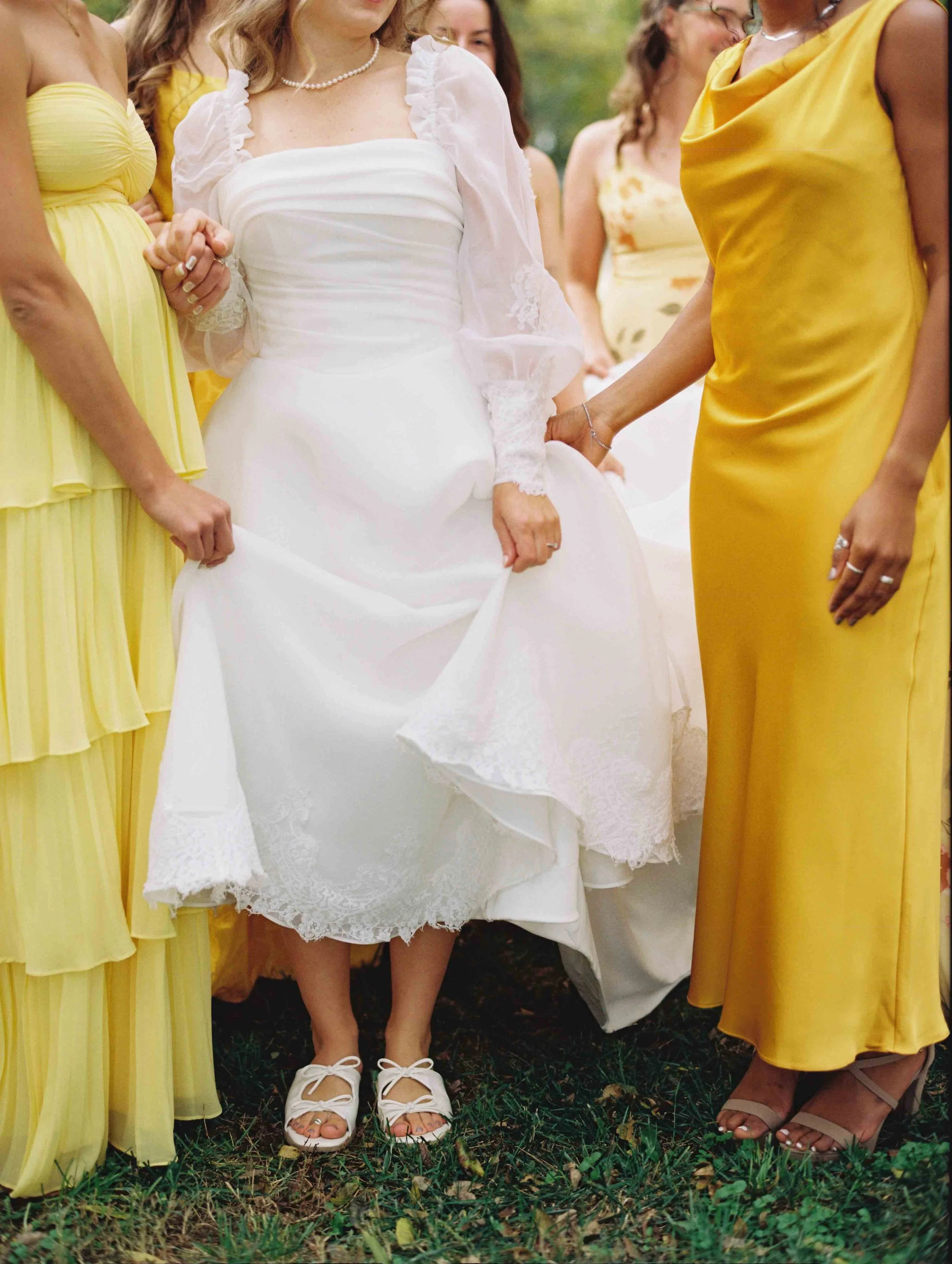 Group of women gathered outdoors, some wearing yellow dresses and a woman in a white dress with lace details, partly visible. The focus is on their lower bodies and feet, standing on grass.