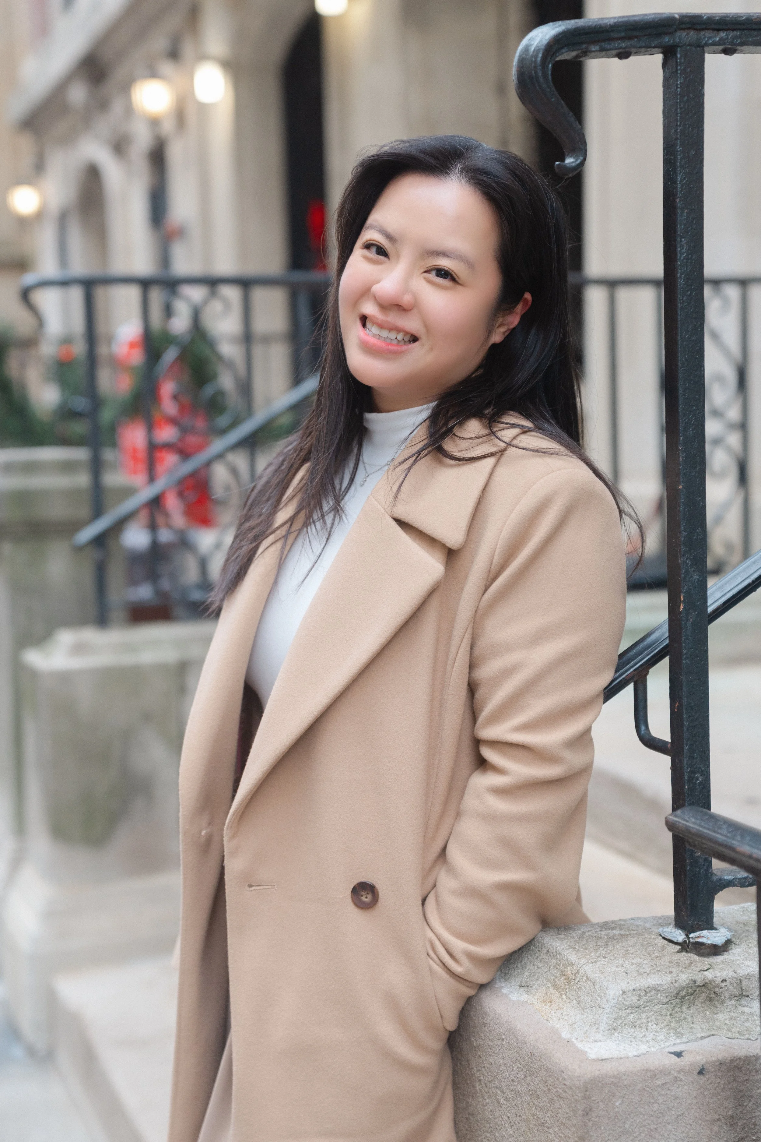 Young woman with long dark hair smiling, wearing a beige coat, standing near a stone ledge with a wrought iron railing in an urban setting.