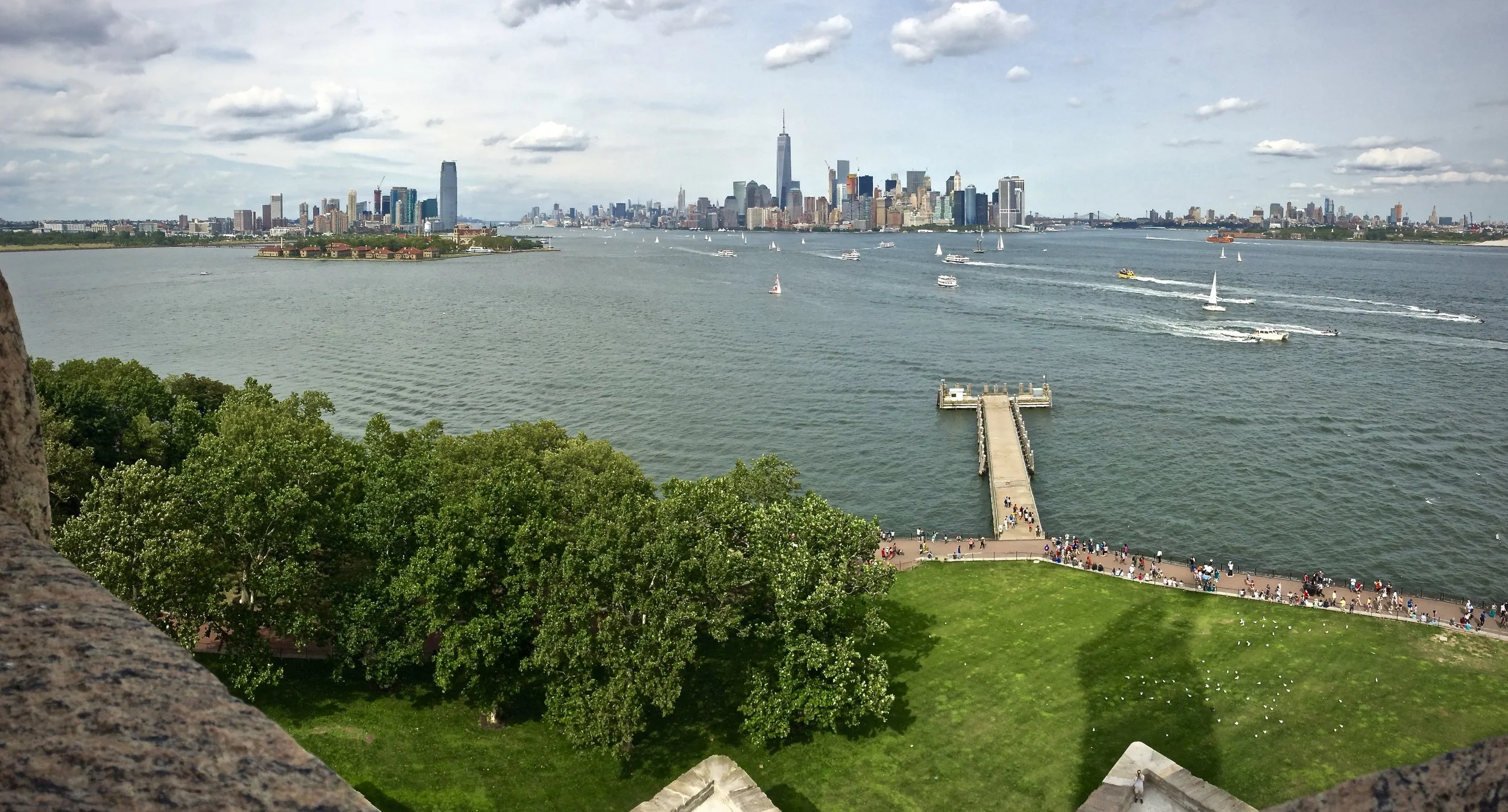 A view of the New York City skyline across a body of water with sailboats, a pier extending into the water, green trees, and people walking on a promenade.