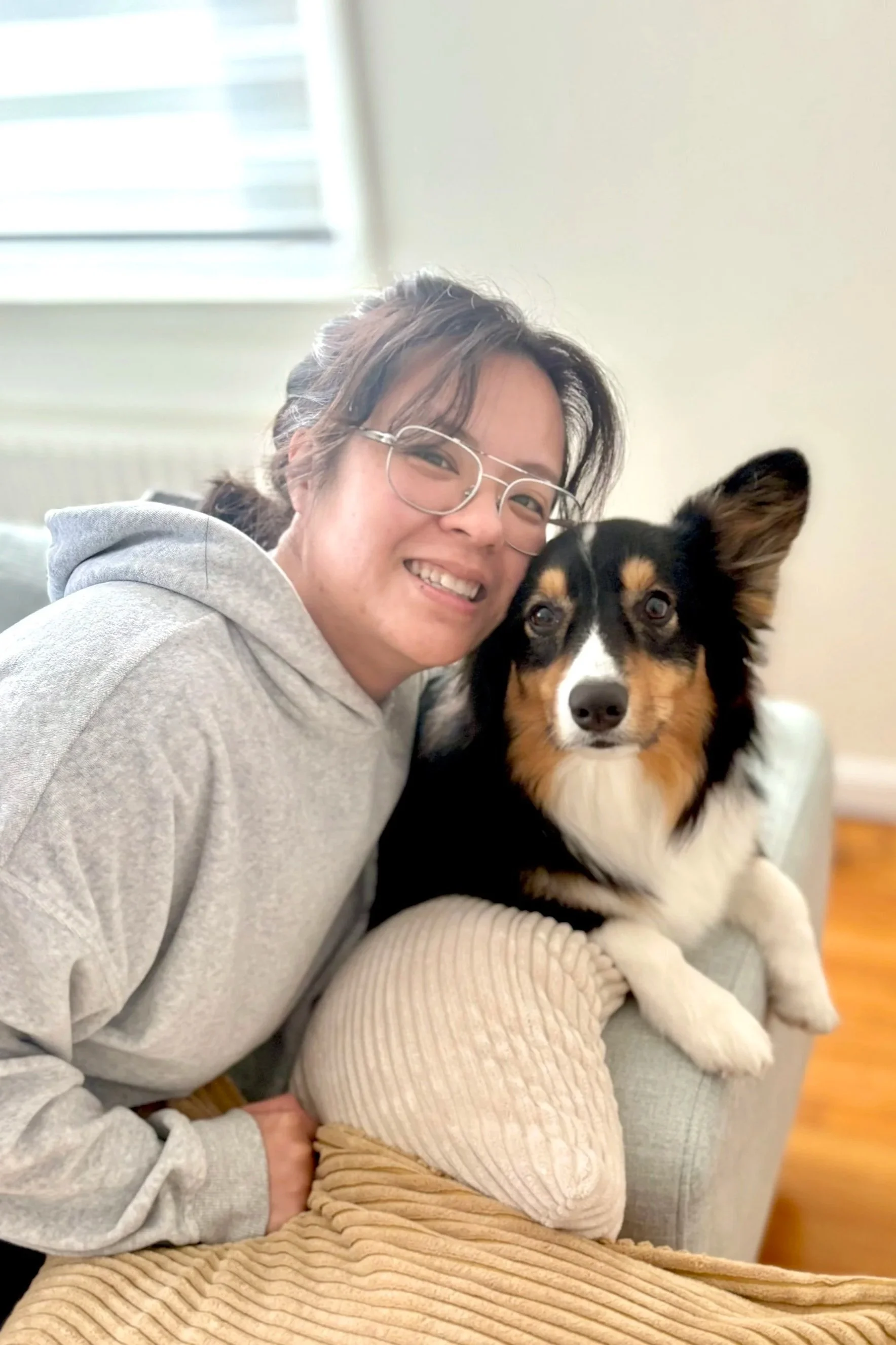 A woman with glasses and a hoodie sitting on a couch, smiling, with a black, white, and tan Australian Shepherd dog sitting on her lap, inside a bright room.