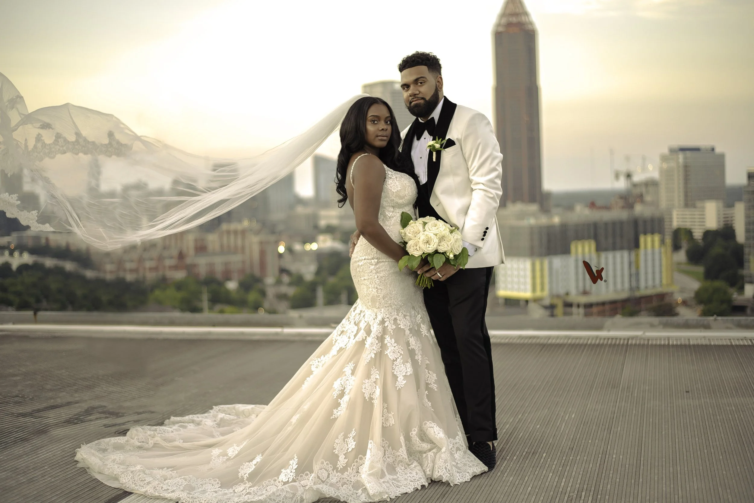 A bride and groom standing on a rooftop with a city skyline in the background during sunset, dressed in wedding attire, with the bride holding a bouquet of white roses
