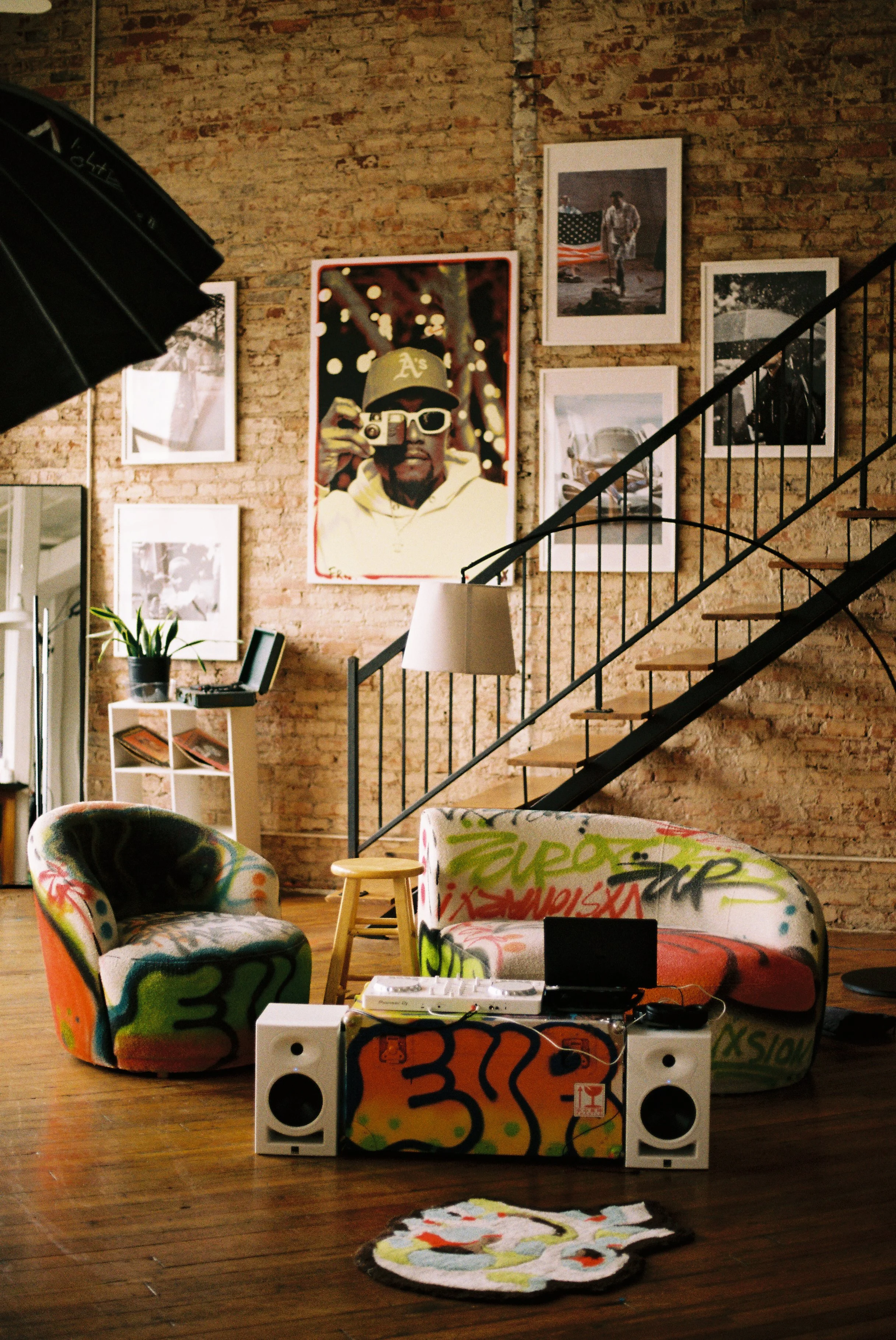Indoor living space with exposed brick walls, graffiti-style furniture, and artwork, including a large portrait of a person wearing a baseball cap and sunglasses holding a camera. Graffiti furniture pieces include a colorful armchair, sofa, and small table, with a stereo system on the floor. Decorated walls feature framed photos and artworks, and stairs with black railings lead up to a second level.