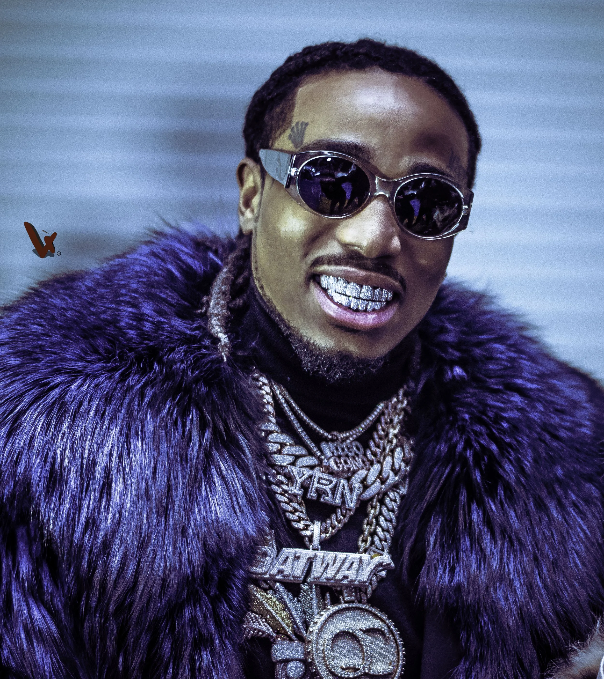 Close-up of a smiling man wearing sunglasses, gold grillz, and a chain with a large pendant, dressed in a dark jacket with a purple or blue fur collar, against a blurred blue background.